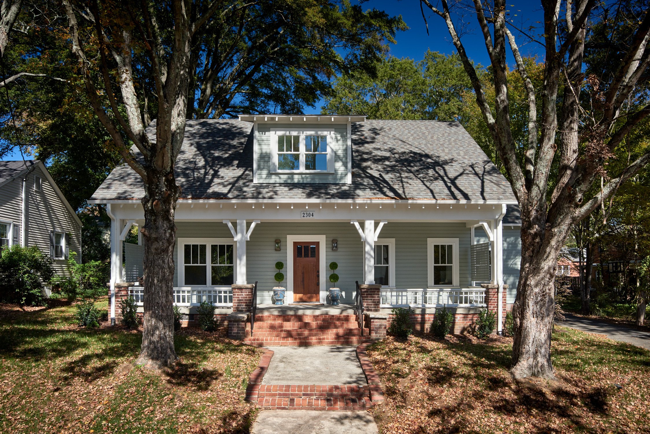 Front view of a two-story house with gray siding, white trim, a gabled roof, and a small front porch with brick steps, wood railing, and two topiary bushes flanking the door. Two large trees frame the house, reflecting fall sunlight and casting shado