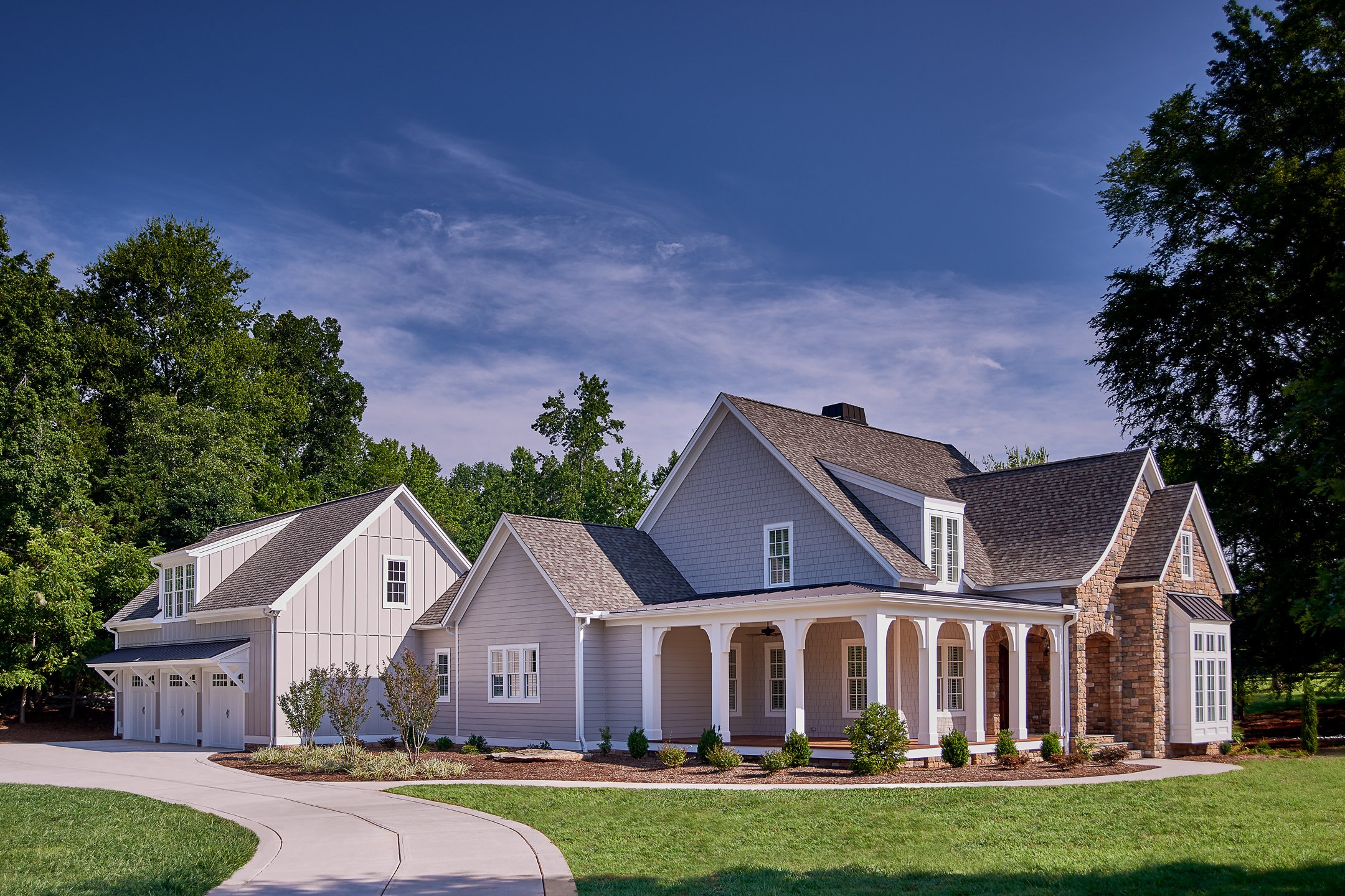 A spacious suburban house with a large front porch, white siding, multiple gables, and a stone chimney, surrounded by green grass and trees under a blue sky.