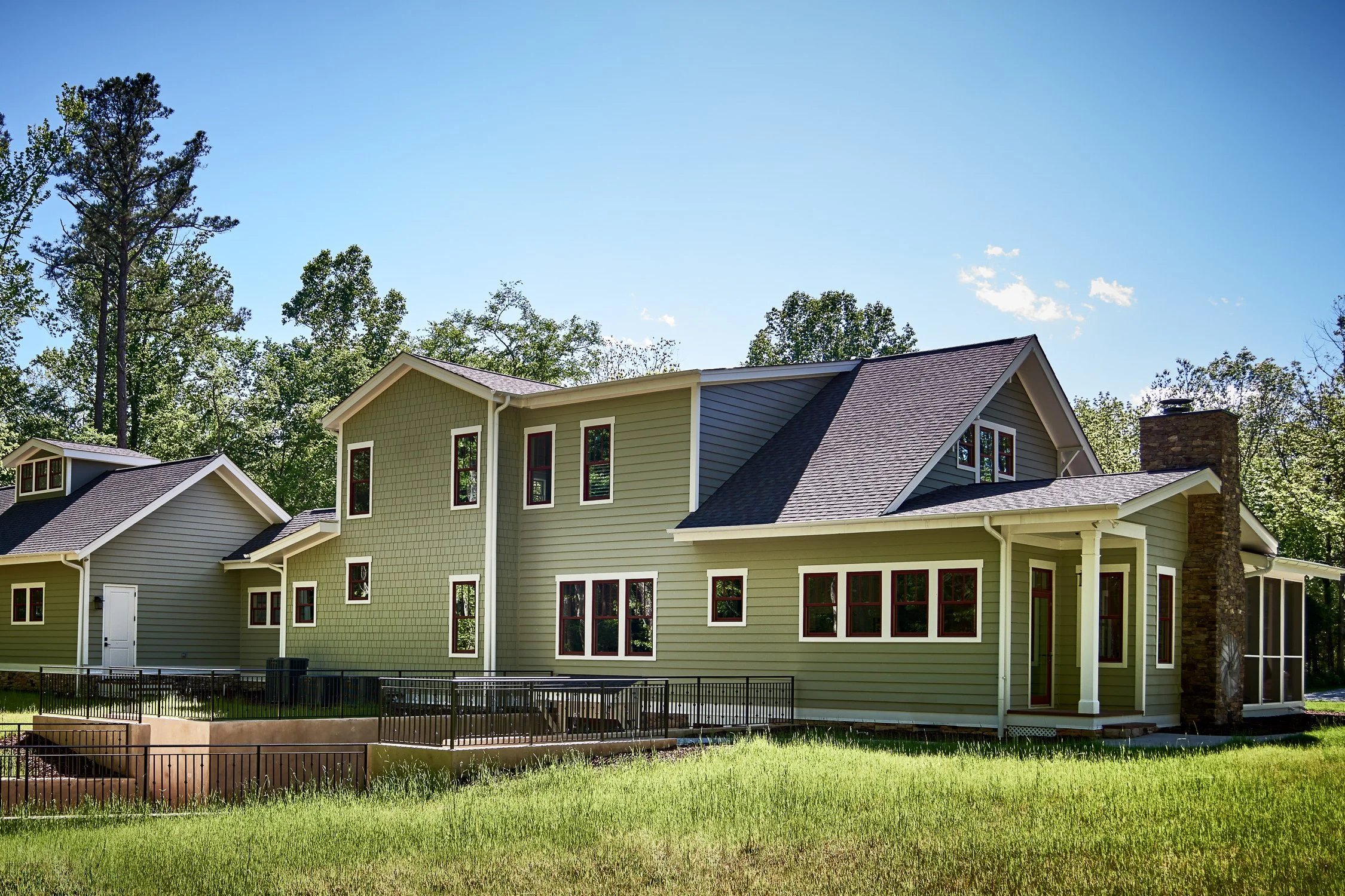 A two-story house with sage green siding, white trim, multiple windows, and a stone chimney, set against a background of tall trees and a bright blue sky.