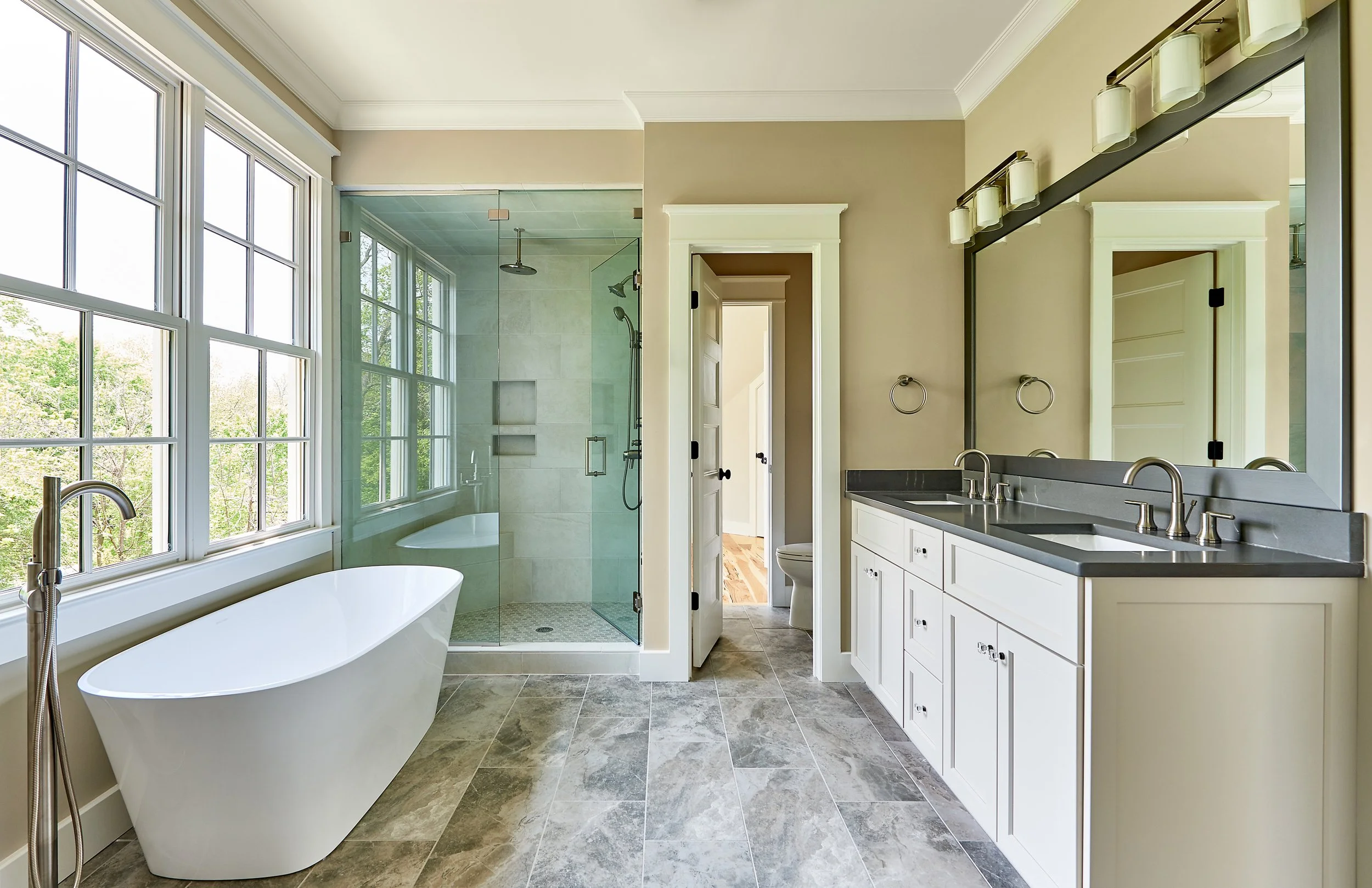 Spacious bathroom with a large soaking tub, a walk-in shower with glass doors, a double sink vanity with a large mirror, and multiple windows overlooking greenery.