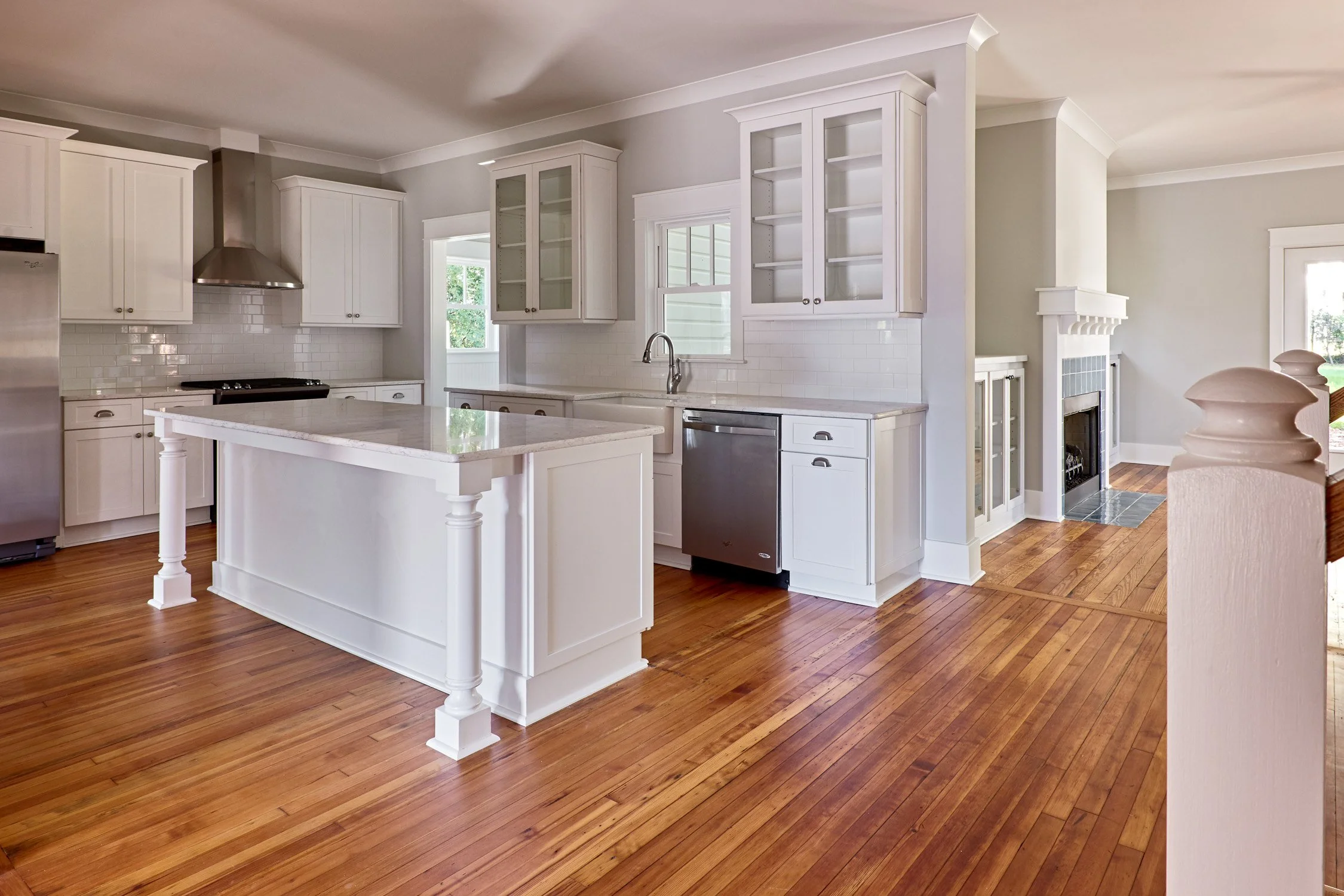 A bright, spacious kitchen with white cabinets, a kitchen island with a marble countertop, hardwood floors, a stainless steel dishwasher, a gas stove, and a fireplace in the adjacent living area.