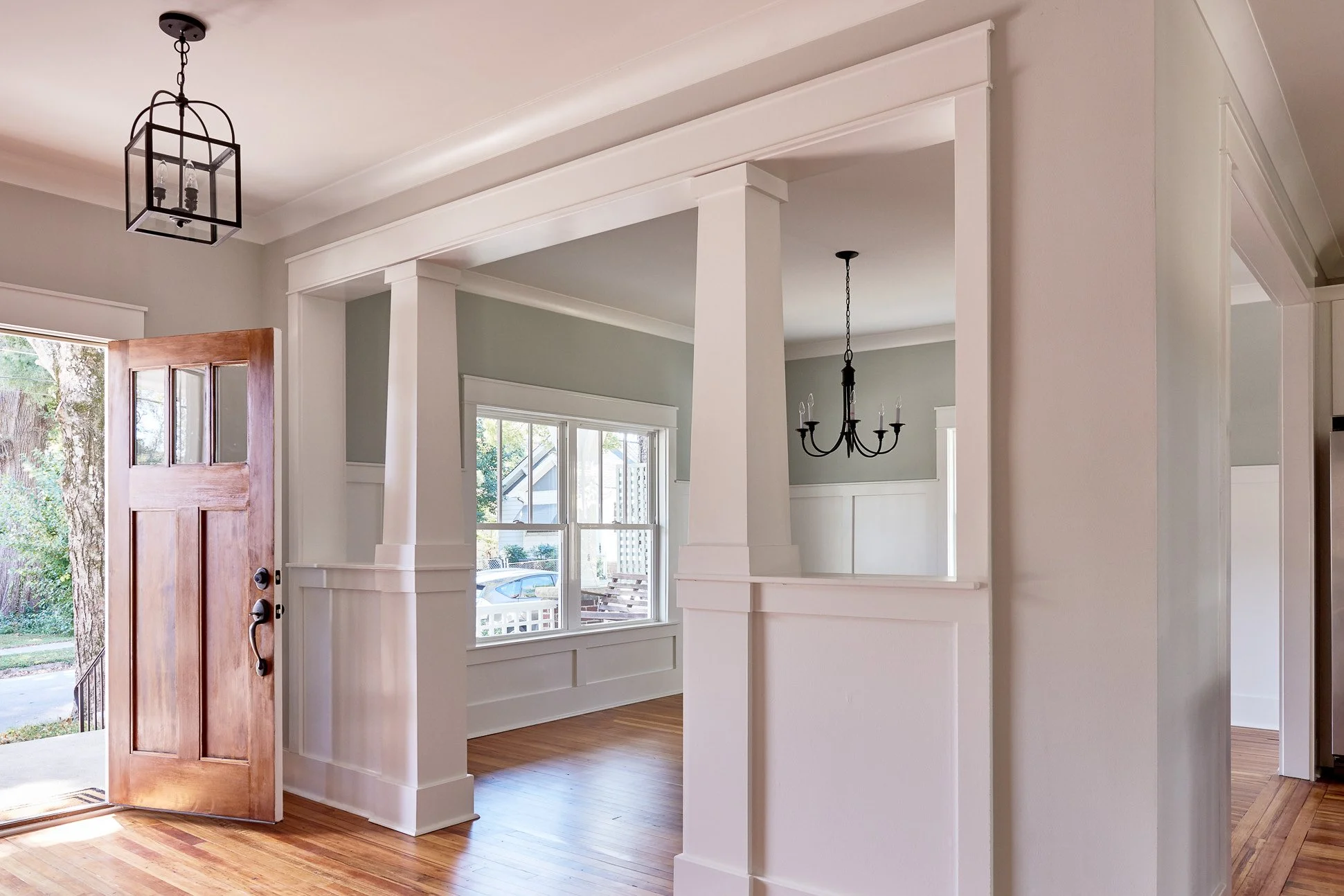 Interior view of a house with hardwood floors, open doorway, and hanging light fixtures. Front door is wooden with window panels, open to outside with visible trees and cars.