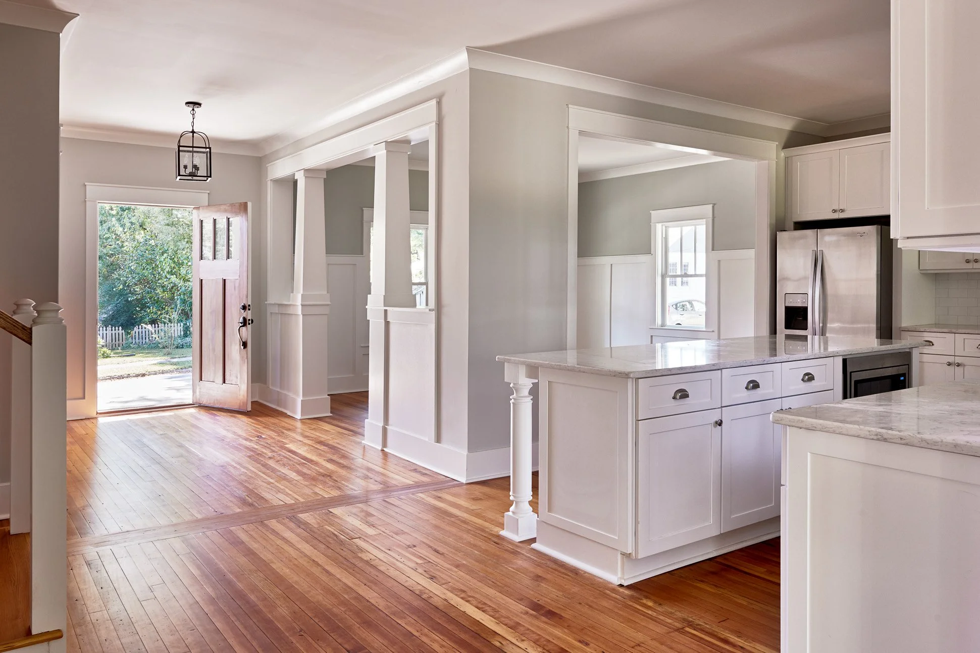 Interior view of a bright, modern kitchen and entryway with hardwood flooring, white cabinets, and stainless steel appliances, including a refrigerator and microwave.