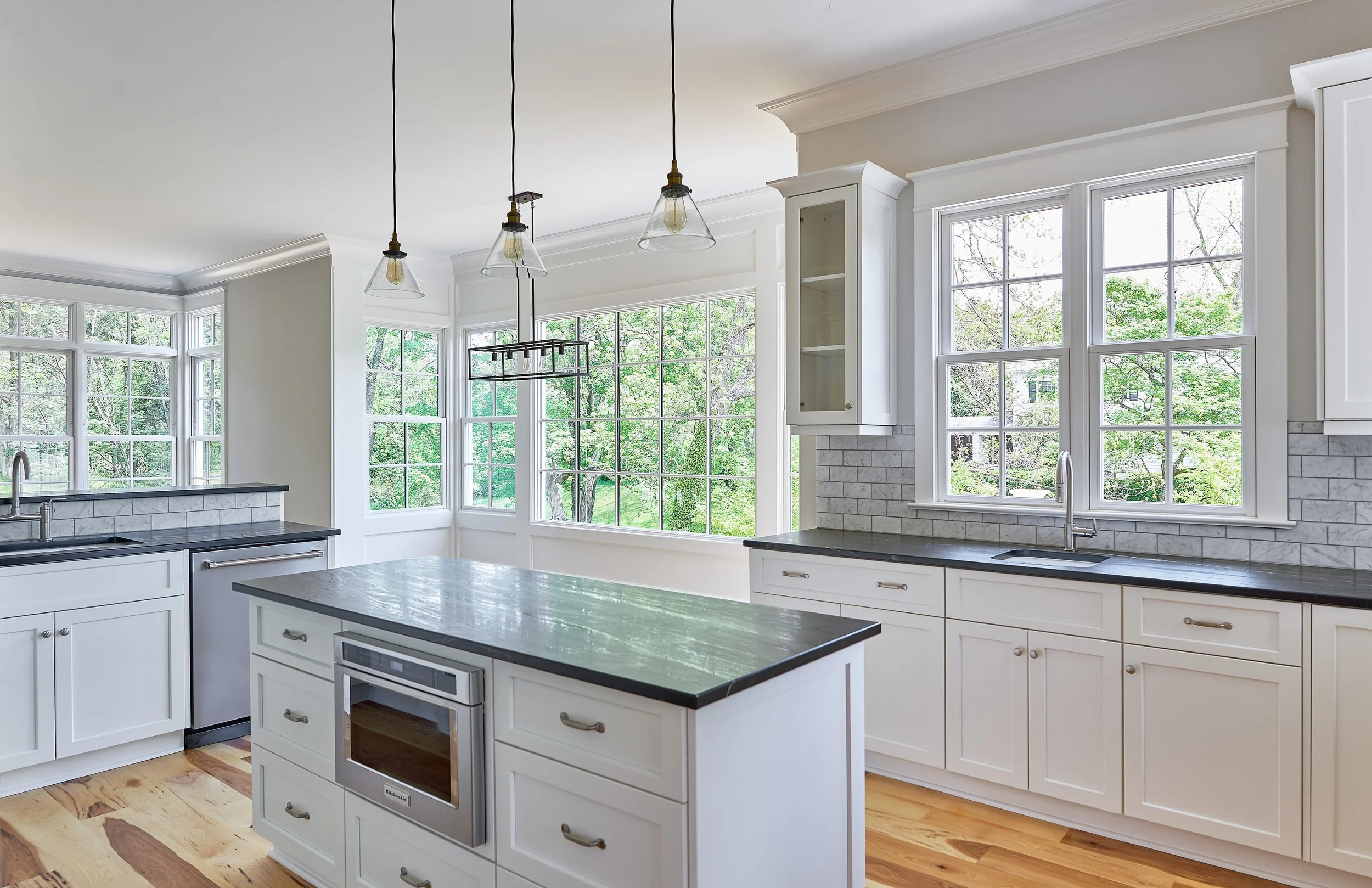 Bright kitchen with white cabinets, large windows with views of green trees, a dark countertop on the island and around the sink, and pendant lights hanging from the ceiling.