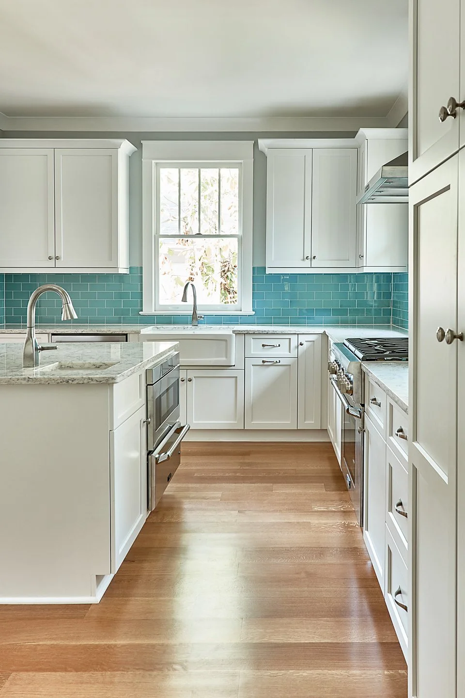 Bright, white kitchen with light wood floor, blue tile backsplash, and stainless steel appliances. Central island features a sink and granite countertop. Window above sink lets in natural light.
