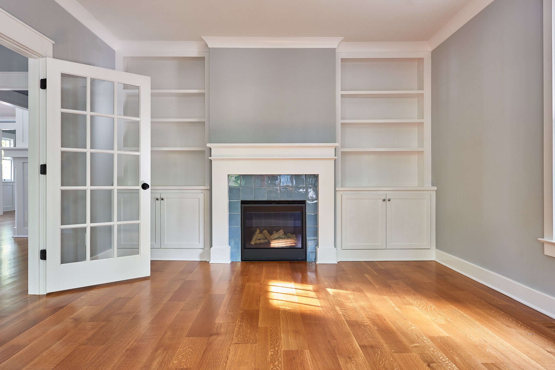 Empty living room with built-in white bookshelves, a white fireplace with a blue tile border, hardwood floor, and light gray walls.