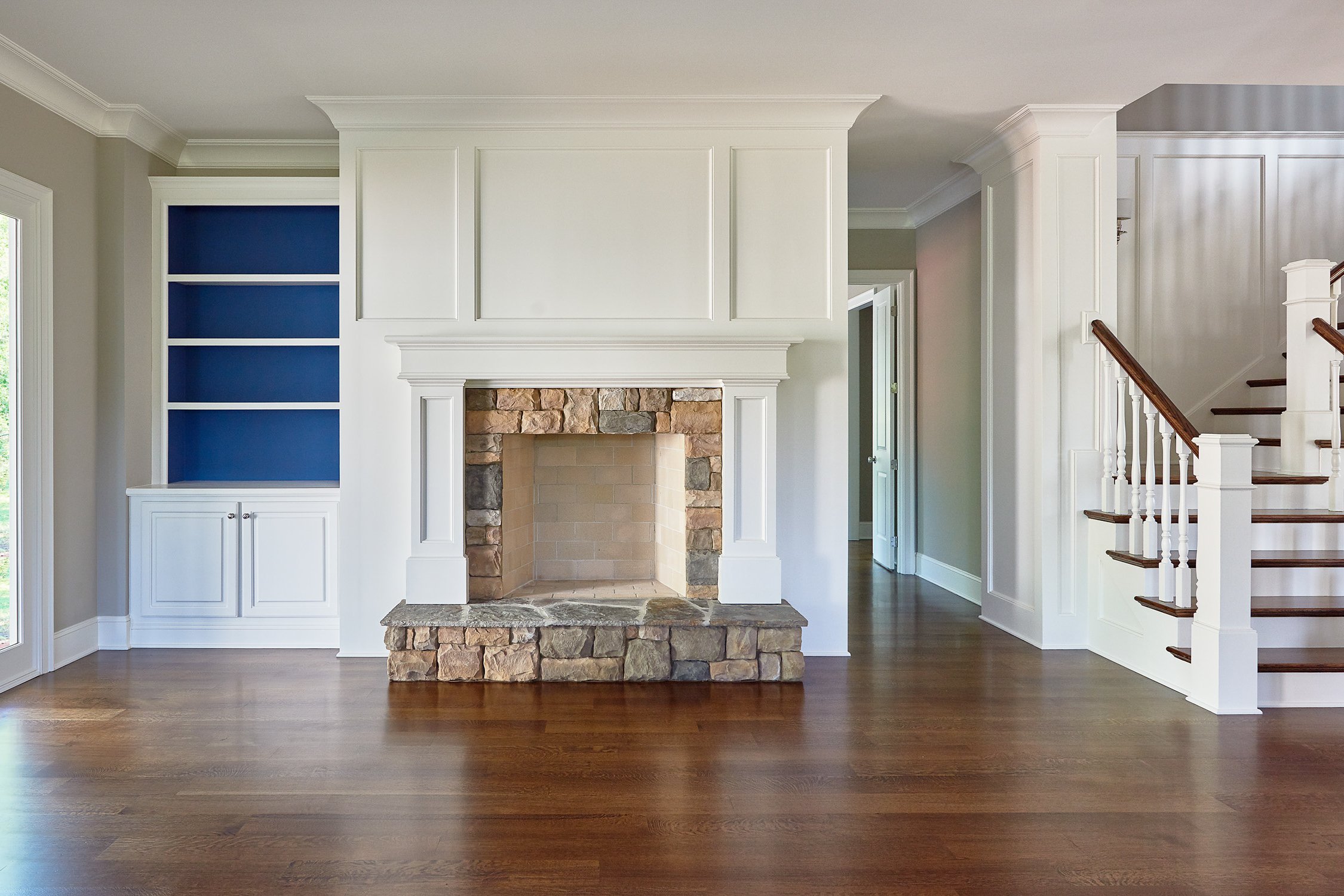 Living room with a fireplace featuring a stone facade, built-in white cabinets, hardwood floors, and a staircase with white risers and dark wood treads.
