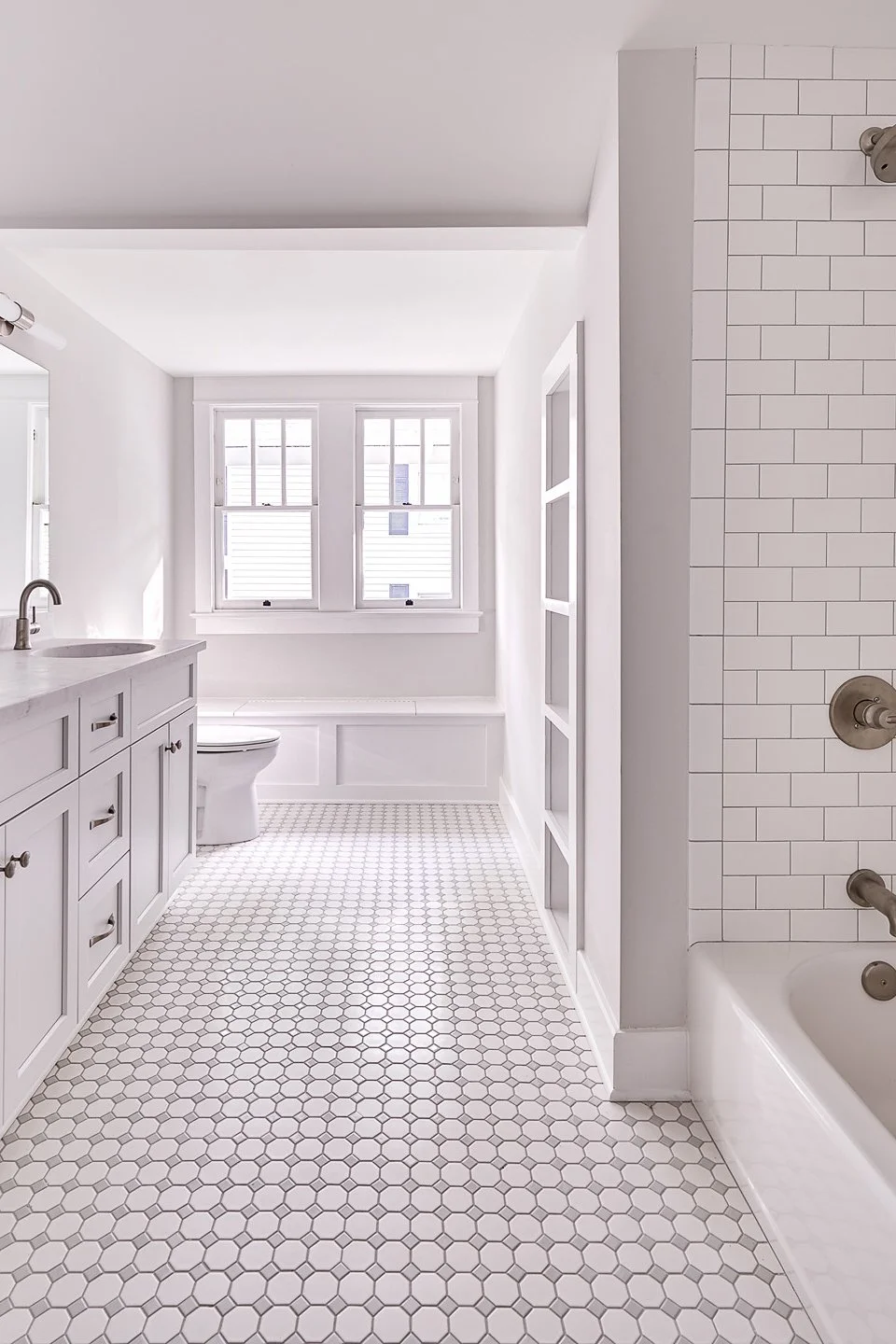 Bright, white bathroom with hexagonal tile floor, vanity with sink, toilet, large windows, and tiled shower area.