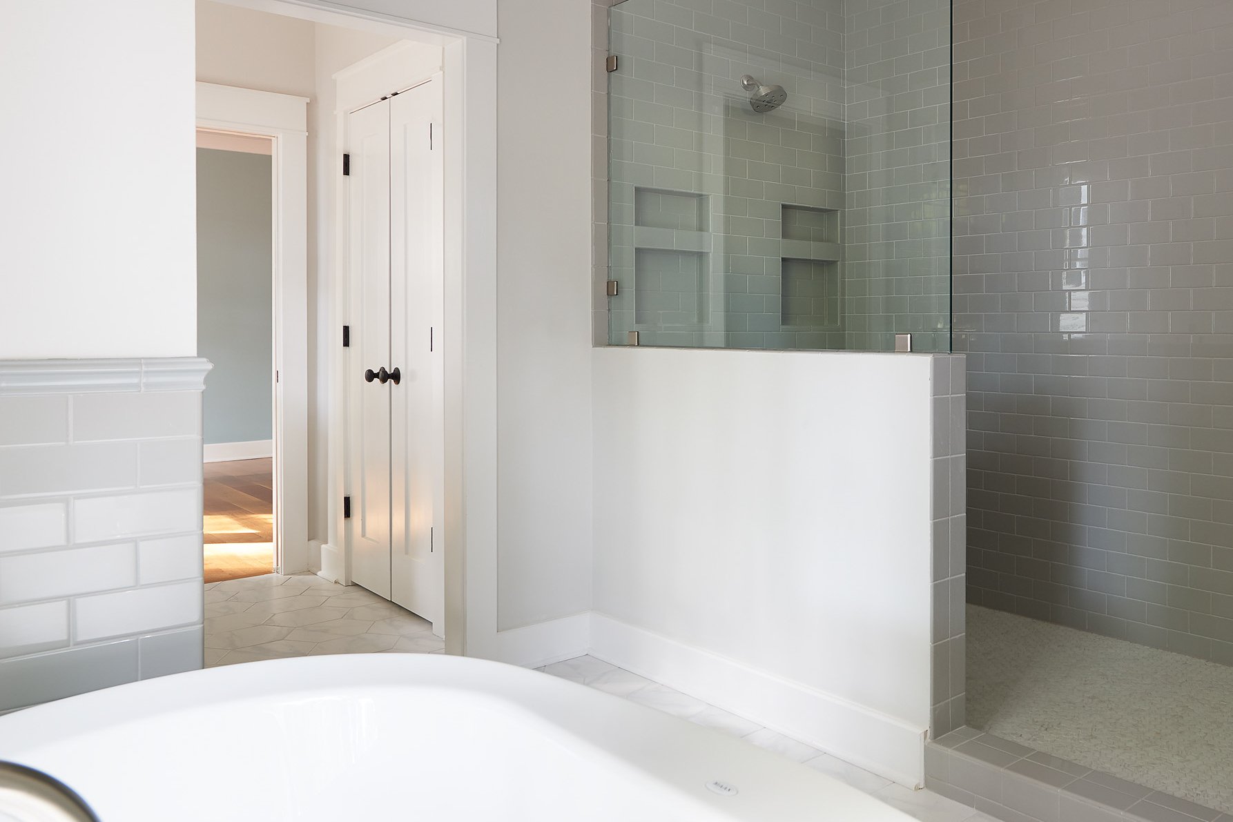 Bathroom with white walls, a walk-in shower with gray tiles and glass enclosure, and a door leading to another room with hardwood flooring.