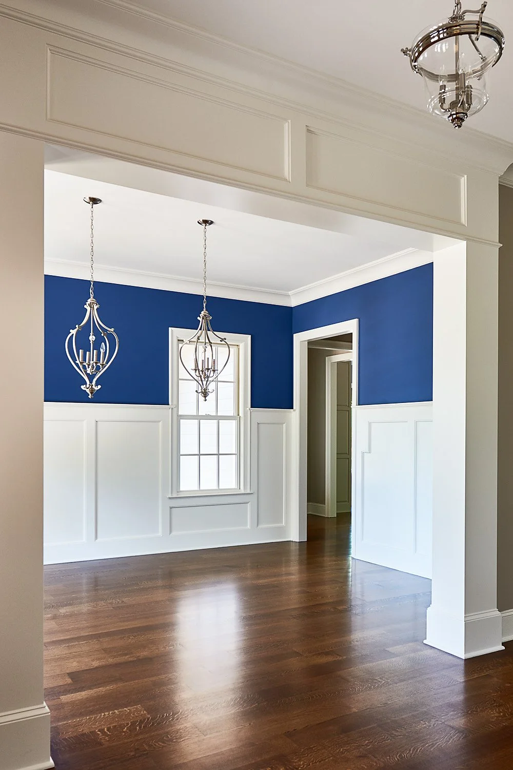 Empty dining room with blue walls, white wainscoting, hardwood floor, a window, and two hanging chandeliers.