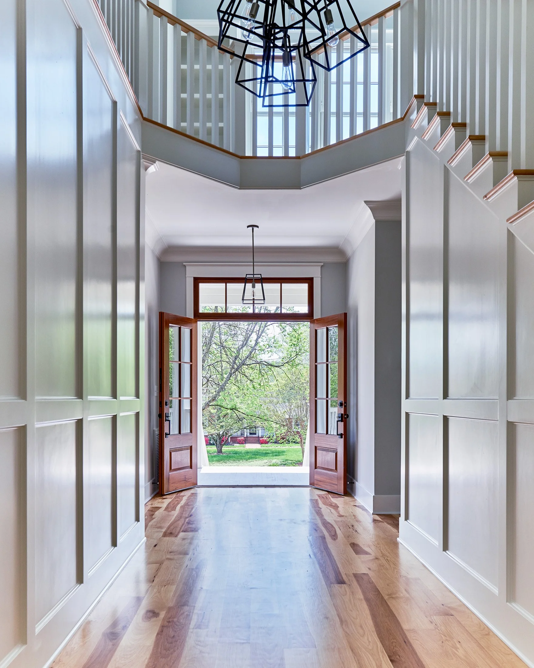 Bright entryway with open wooden front doors leading outside to a green yard with trees, white walls, wooden trim, and modern geometric chandelier.