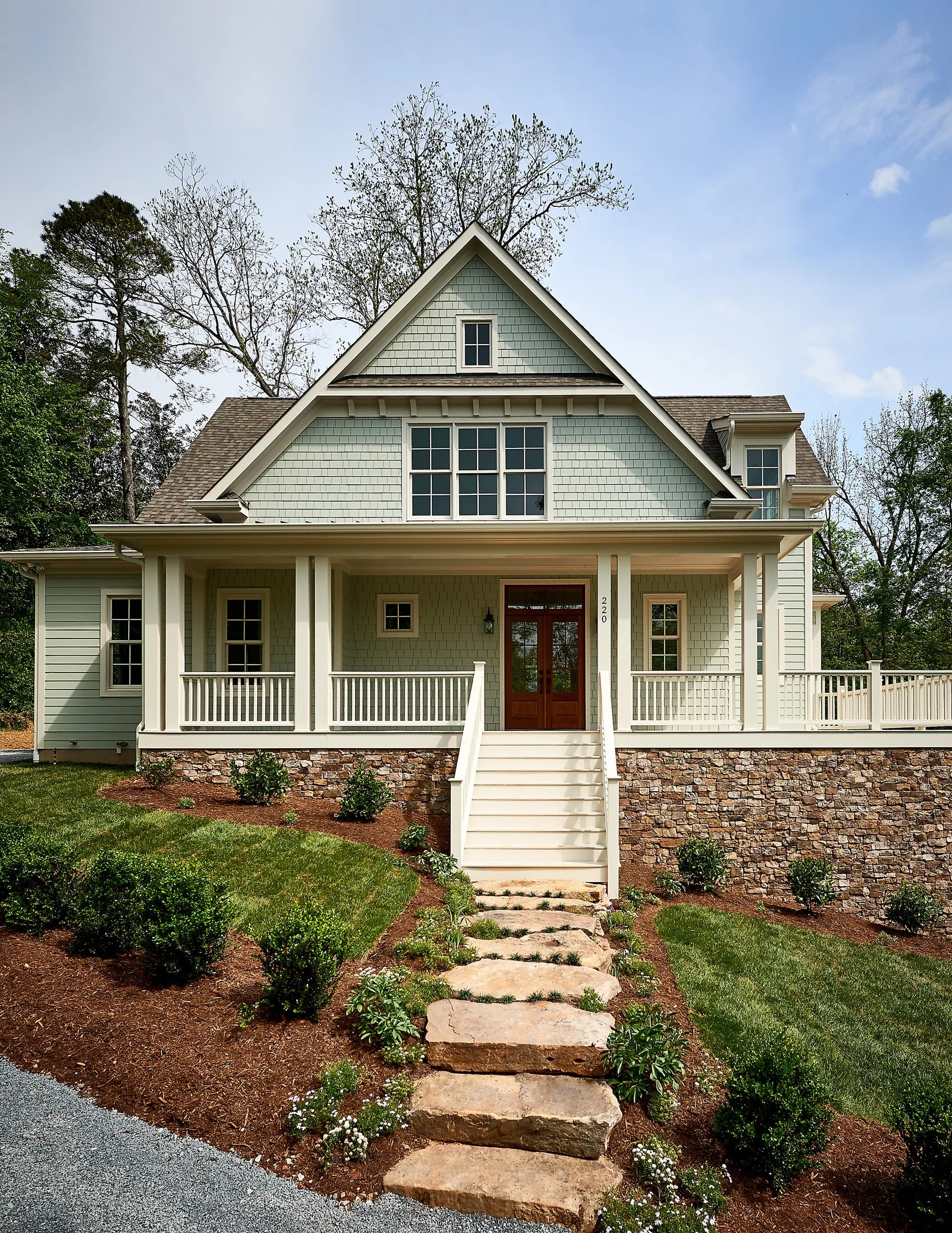 A two-story house with a front porch, stone stairs, green lawn, and landscaped garden, painted in light colors with a gabled roof.