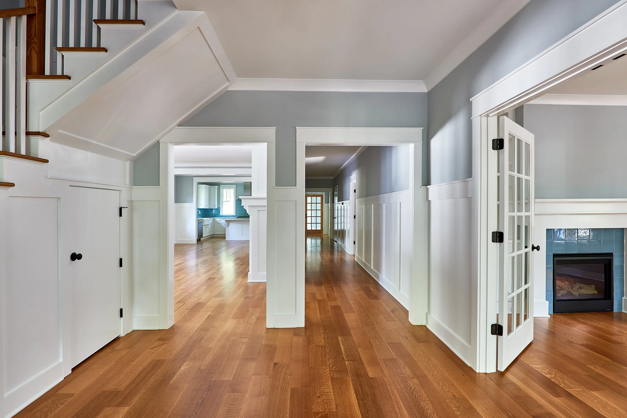 An interior view of a house with wooden floors, gray walls, and white trim. There is a staircase on the left, an open doorway to a kitchen in the background, and a hallway leading further inside. To the right, there is a closed glass-paneled door lea