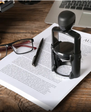 A stapler, a pen, and eyeglasses resting on a printed document on a wooden desk, with a laptop in the background.