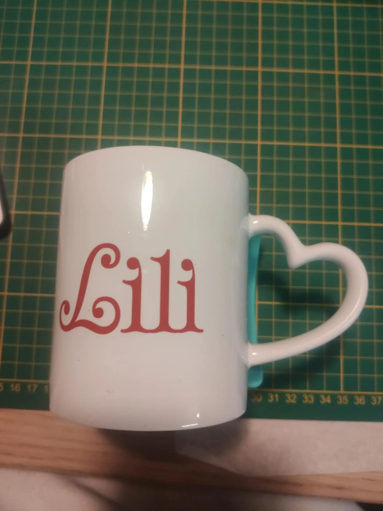 Tasse blanche avec le mot 'Lili' écrit en rouge, posé sur une table avec un tapis de découpe vert en arrière-plan.