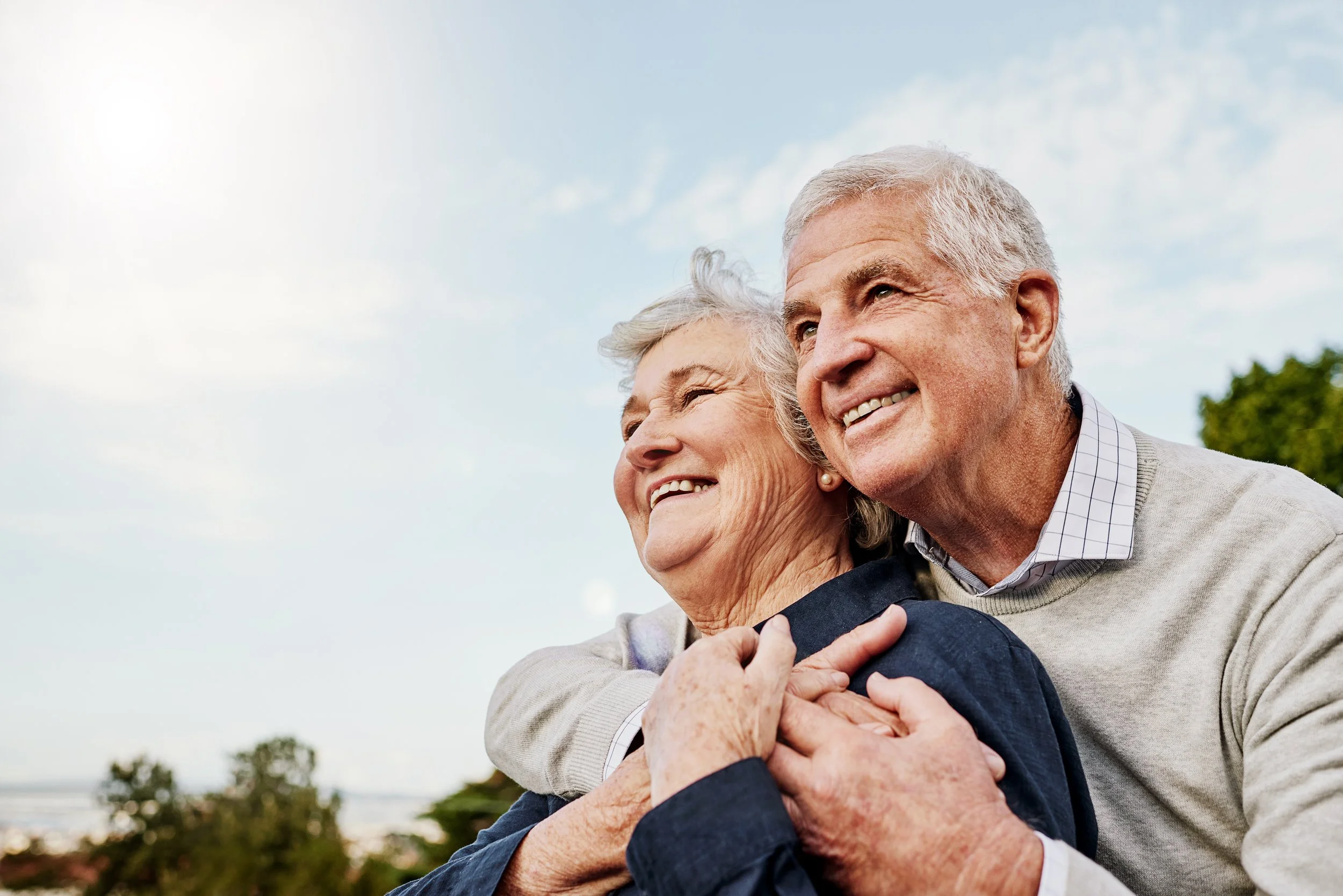 An elderly couple smiling and hugging outdoors under a blue sky with clouds.