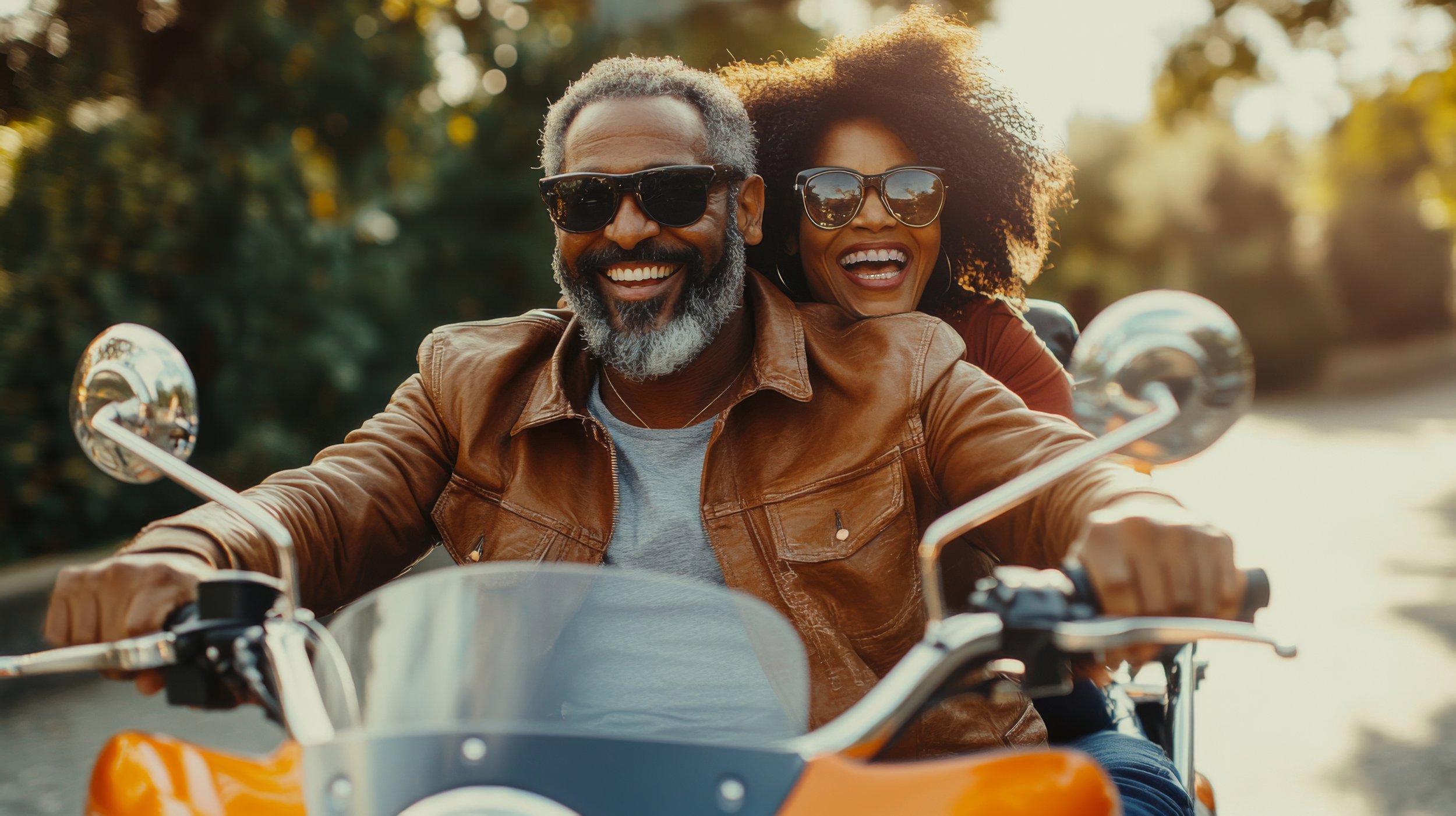 A happy older man and woman riding a motorcycle together outdoors on a sunny day, both wearing sunglasses and smiling.