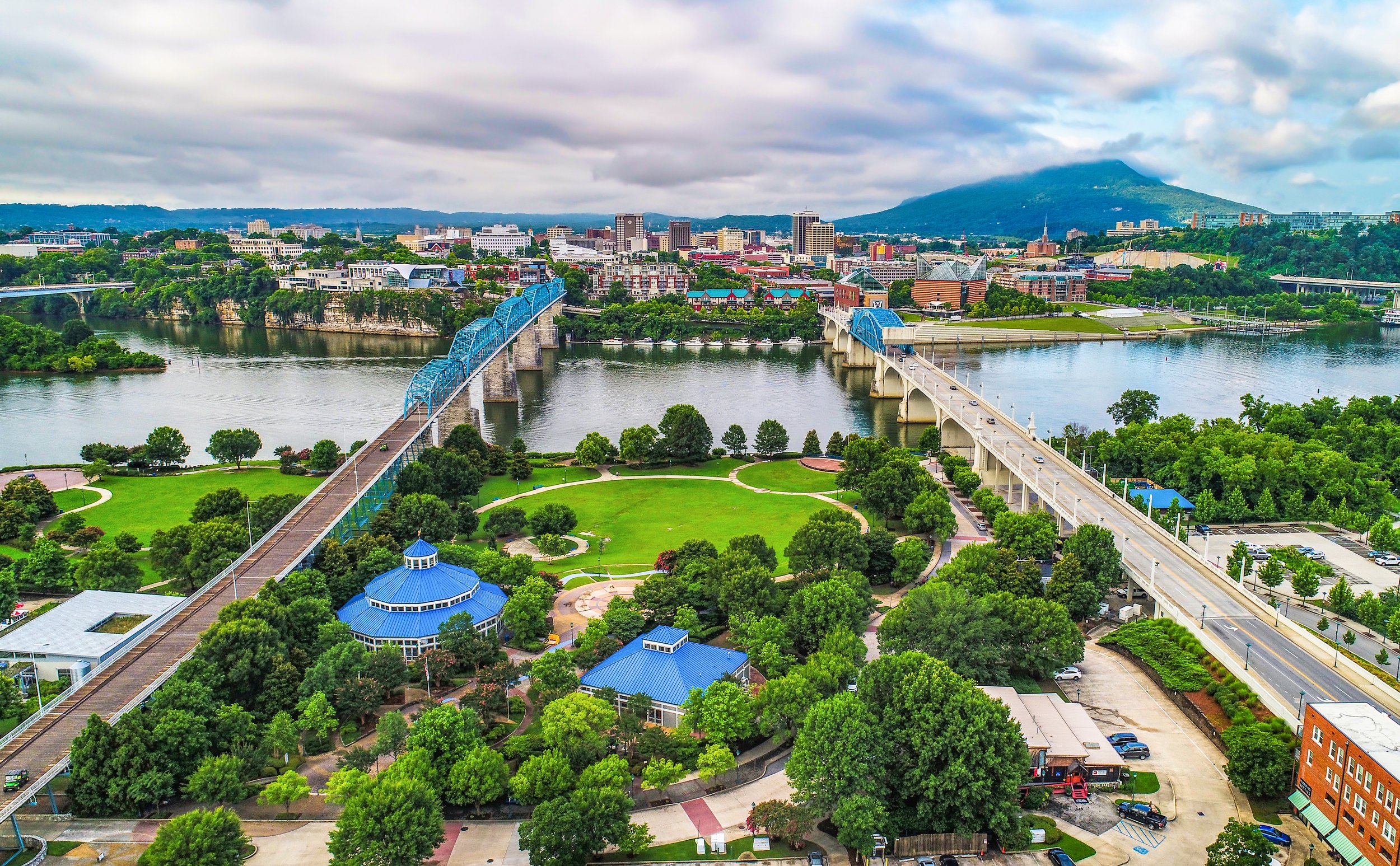 A panoramic view of a cityscape with a river, a bridge, green park areas, and a mountain in the background.