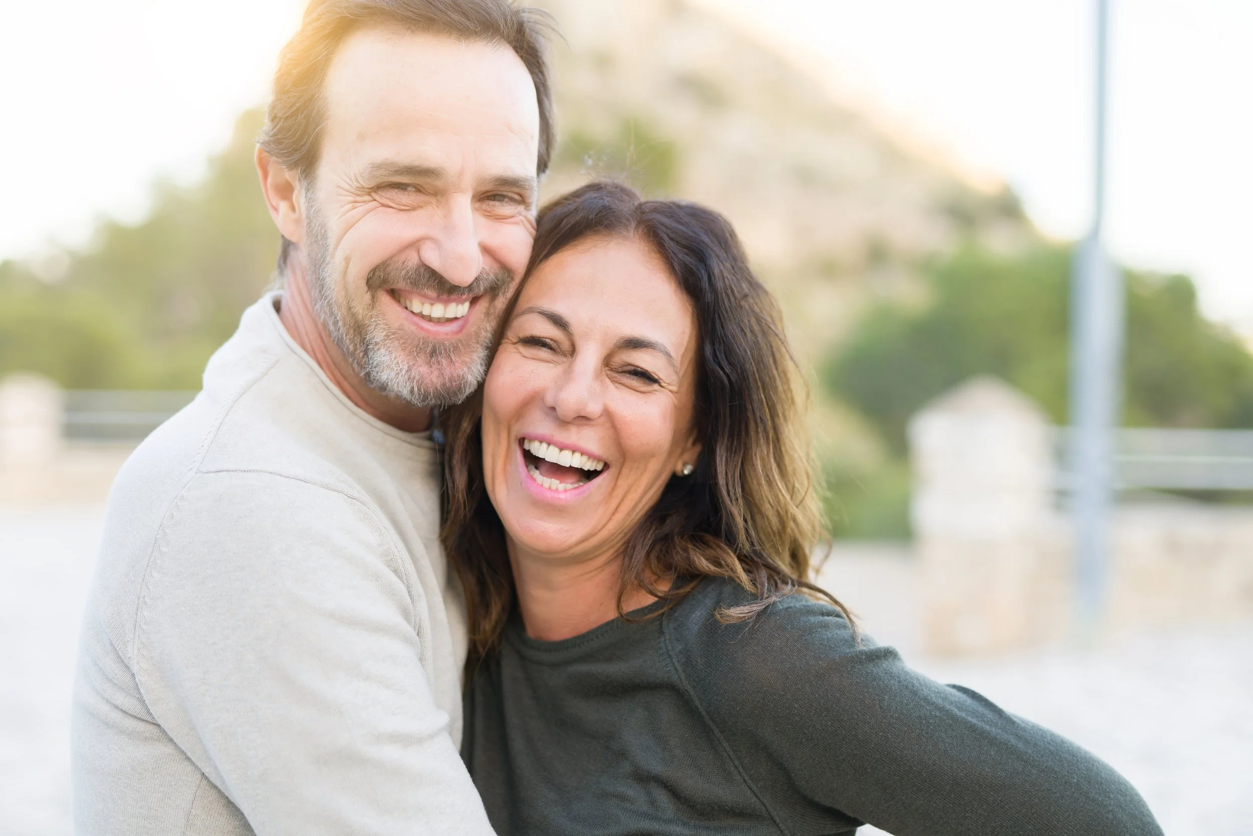 A smiling middle-aged man and woman hugging outdoors in bright sunlight, green trees in the background.