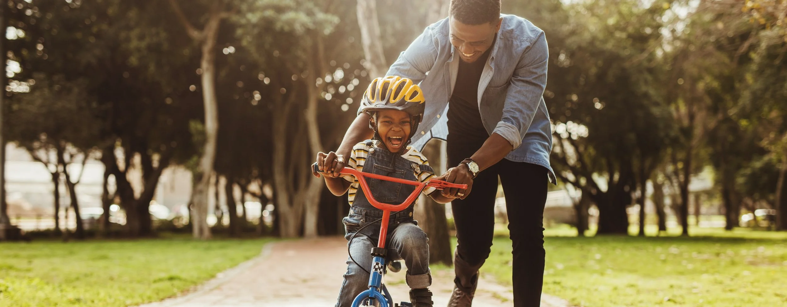 A joyful child wearing a yellow helmet and striped shirt learns to ride a bicycle with the help of an adult in a park with trees and sunlight.