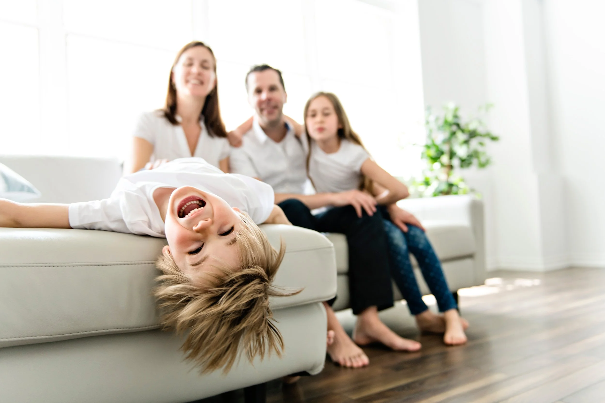 Child lying upside down on sofa, laughing with family of four sitting behind him, in brightly lit living room.