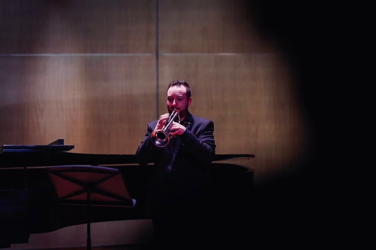 A man in a dark suit playing a trumpet in front of a grand piano, with a music stand nearby, against a plain brown wall.