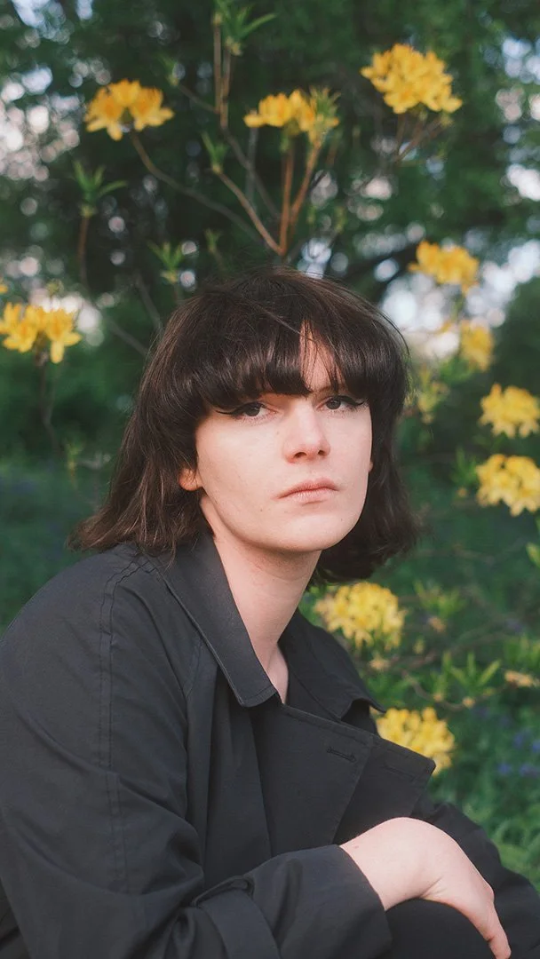 A person with dark, wavy hair and bangs wearing a black jacket, sitting outdoors in front of a bush with yellow flowers.