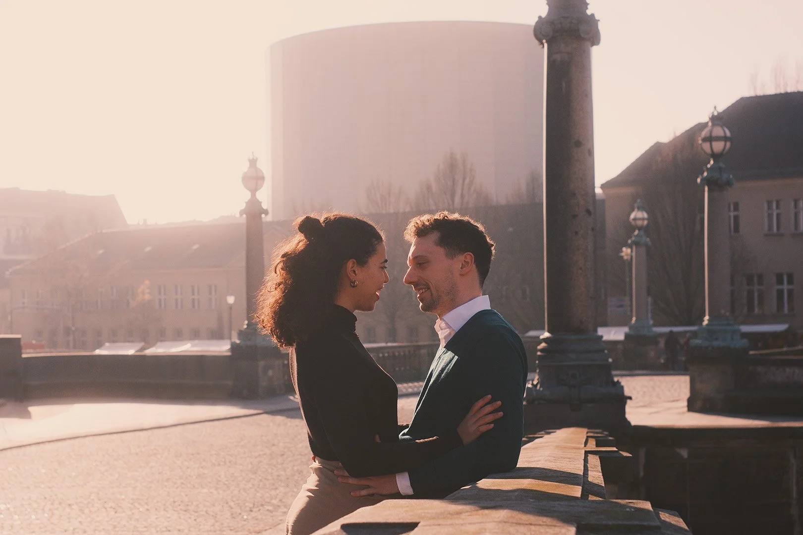 A young couple sharing a tender moment, smiling at each other, on a city street during sunset, with historic lamp posts and a modern building in the background.