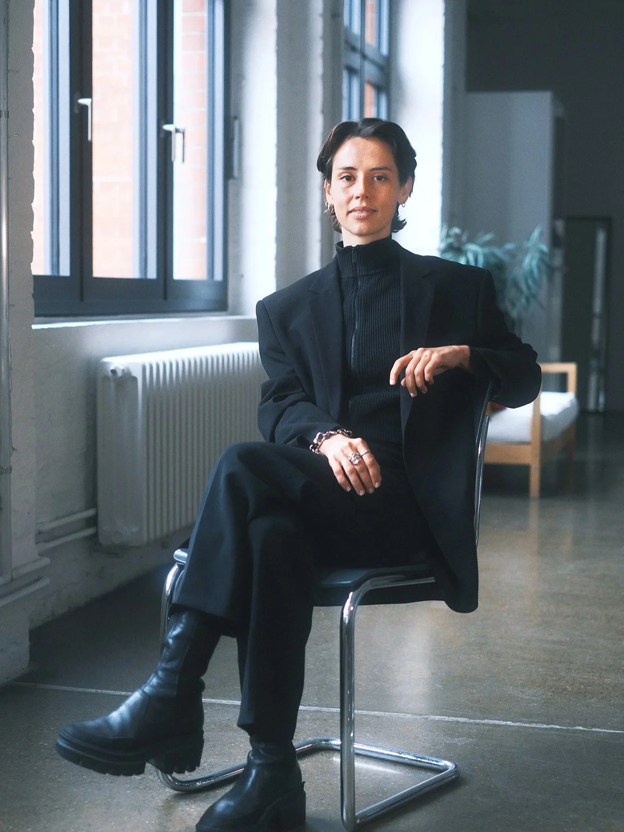 A woman with short dark hair, dressed in a black blazer and pants, sitting on a chair in a modern room with large windows, a radiator, and a potted plant in the background.