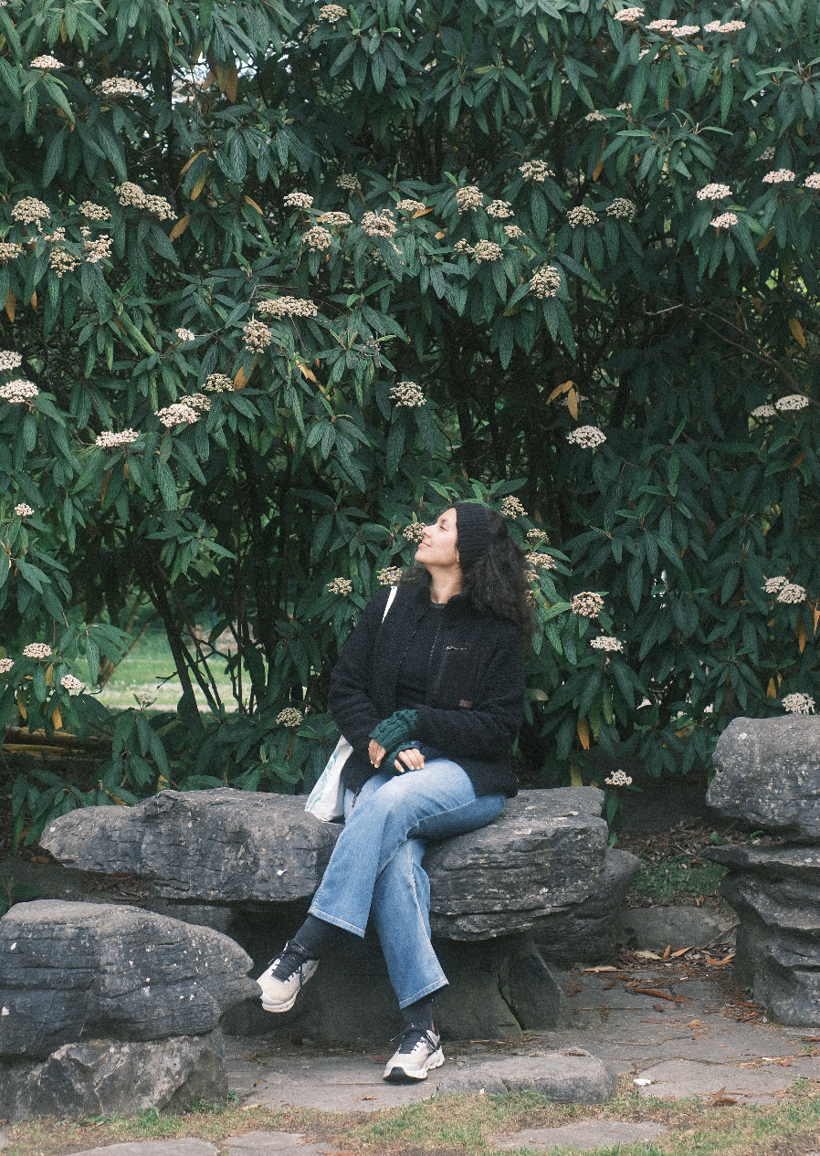 A woman sitting on a large rock in front of a large shrub with white flowers, wearing a black jacket, blue jeans, sneakers, and a black beanie, with a white bag over her shoulder, looking up at the bushes.