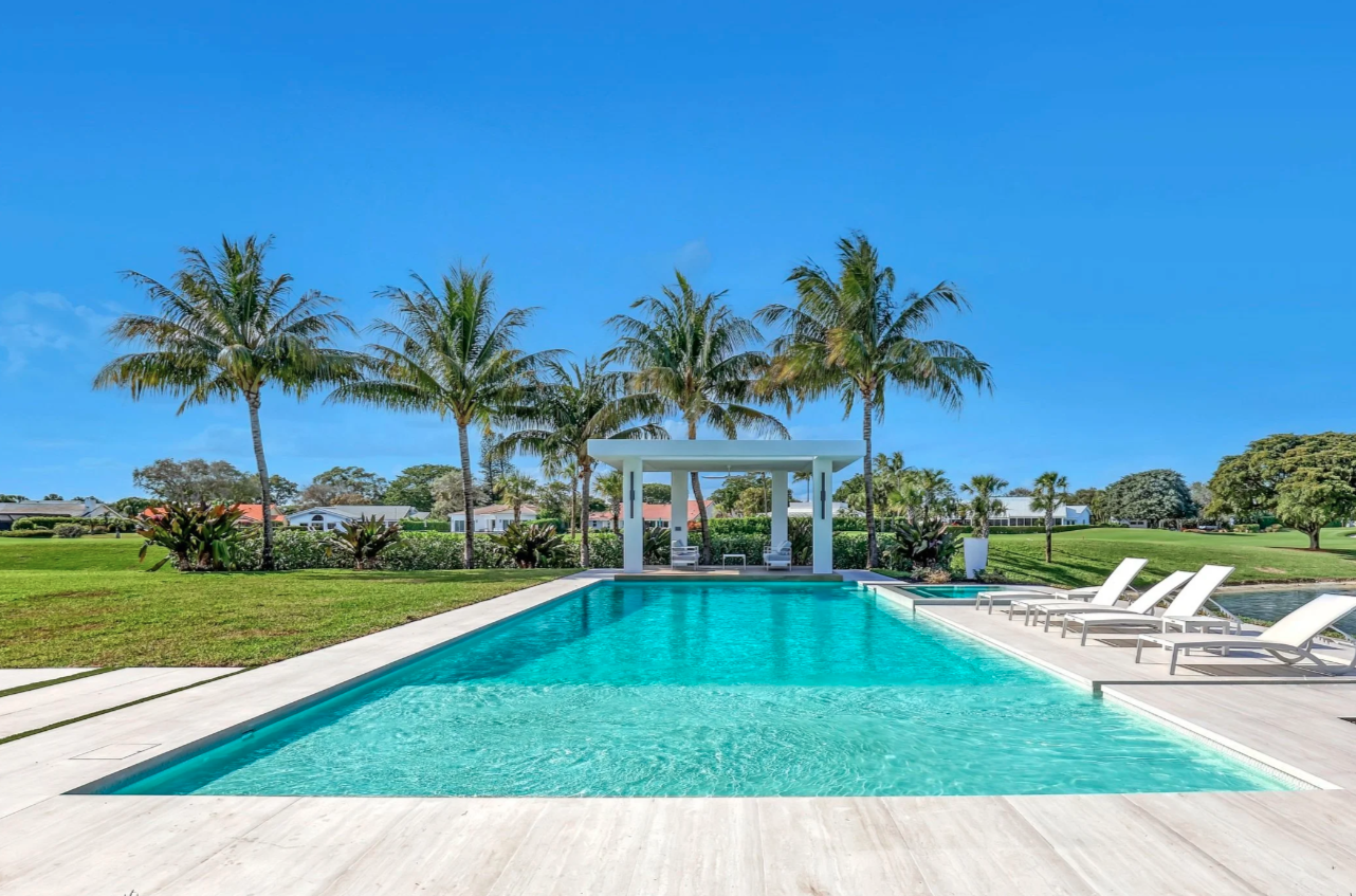 Hang 10 Pools Built Luxury outdoor swimming pool with white lounge chairs on a white deck, surrounded by palm trees and lush green grass under a clear blue sky.