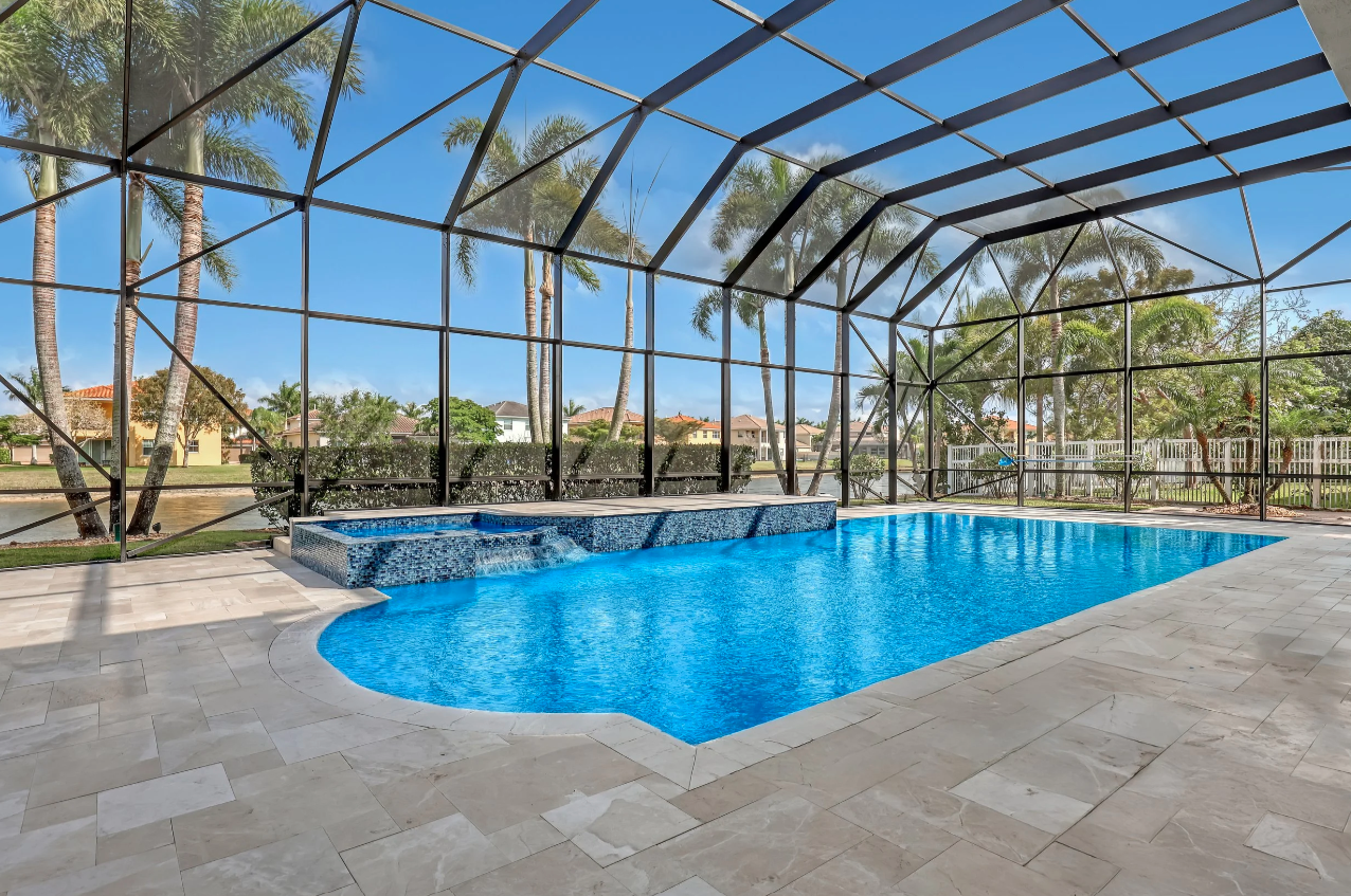 Hang 10 Pools built. Enclosed backyard pool with blue water, tiled hot tub, and tropical trees outside screened-in enclosure under a blue sky.