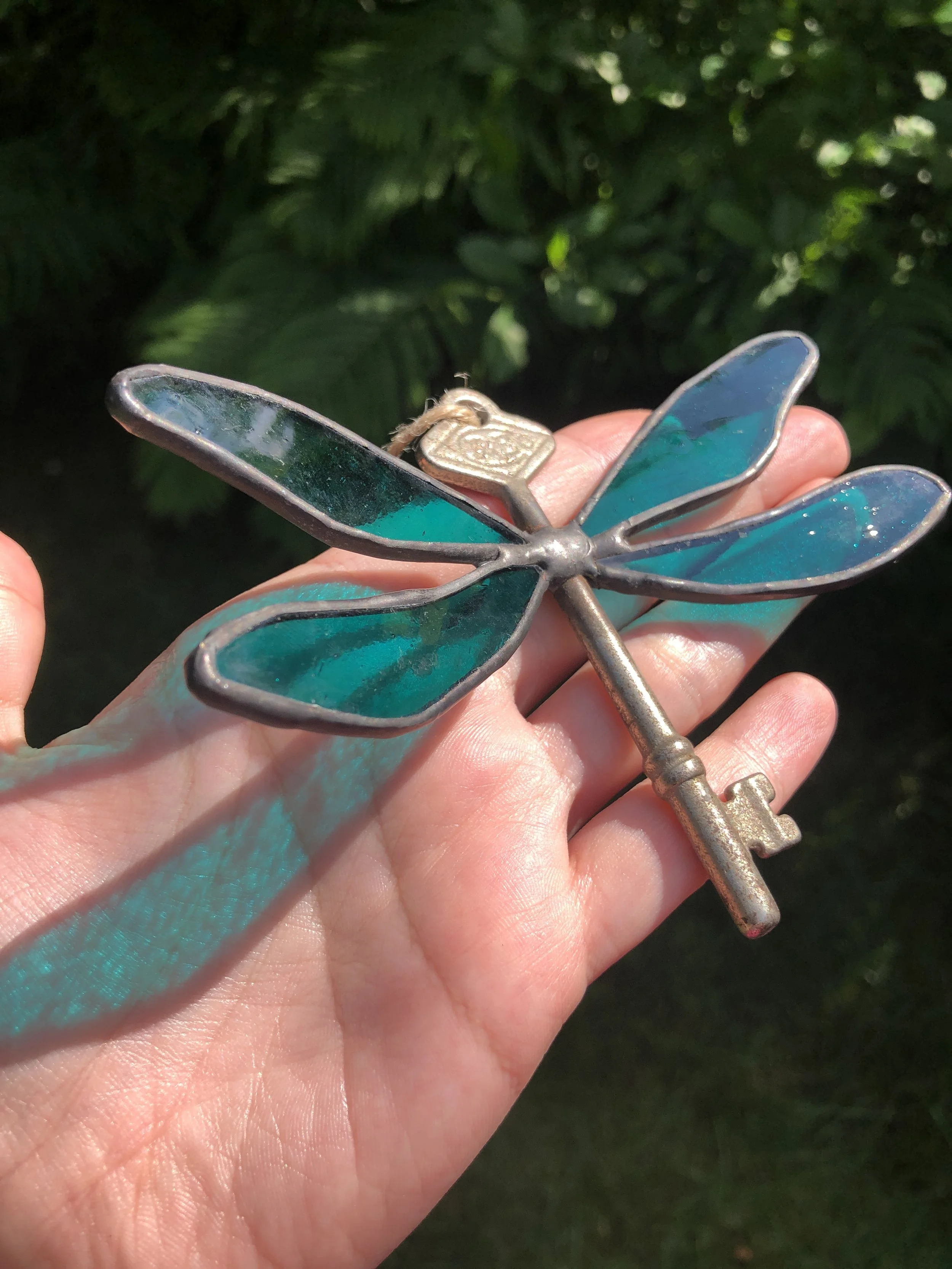 A hand holding a decorative metal dragonfly with blue-tinted wings and an ornate key-shaped tail, outdoors with greenery in the background.