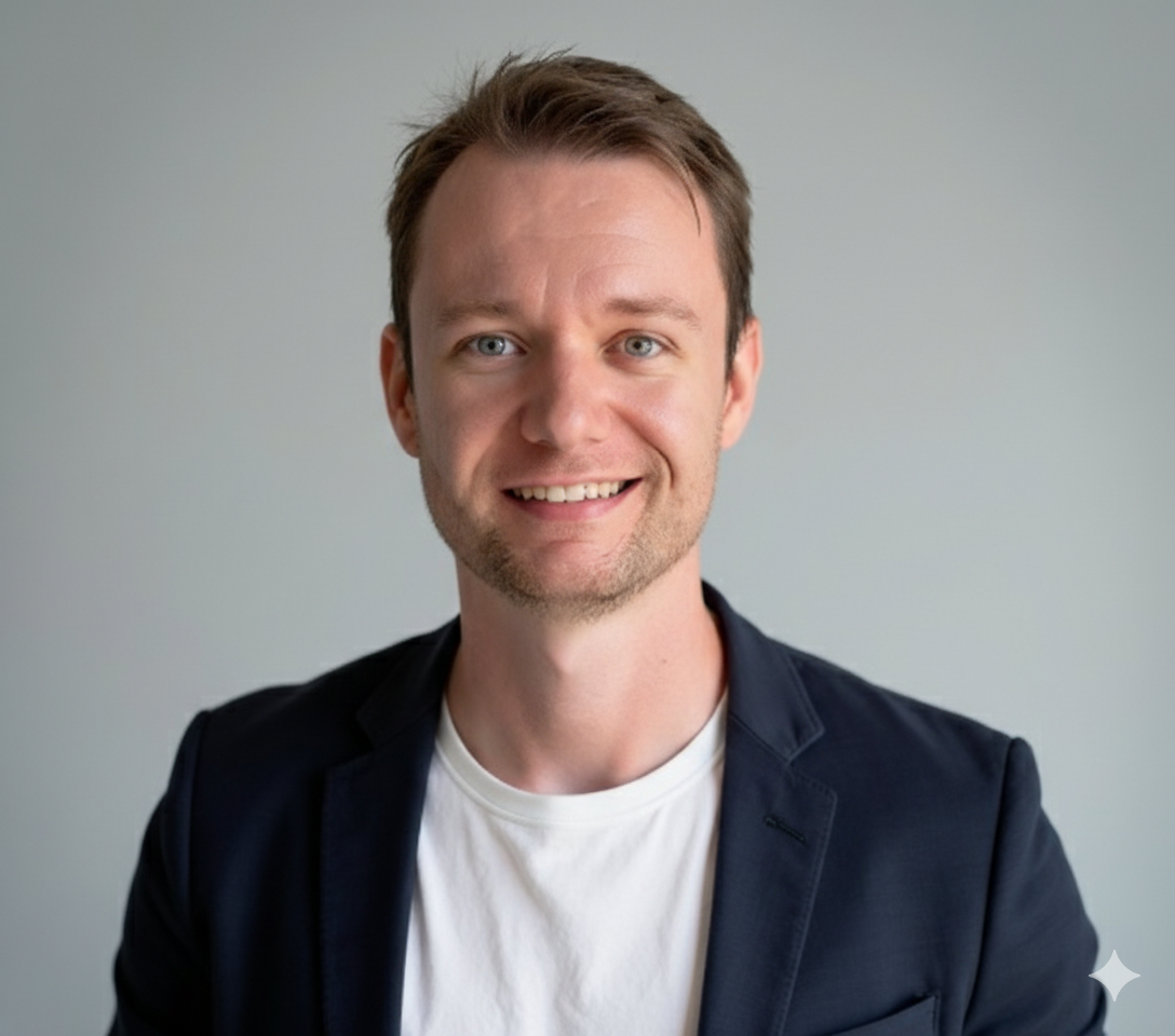 Portrait of a smiling man with short brown hair, wearing a white T-shirt and dark blazer, standing in front of a plain light gray background.