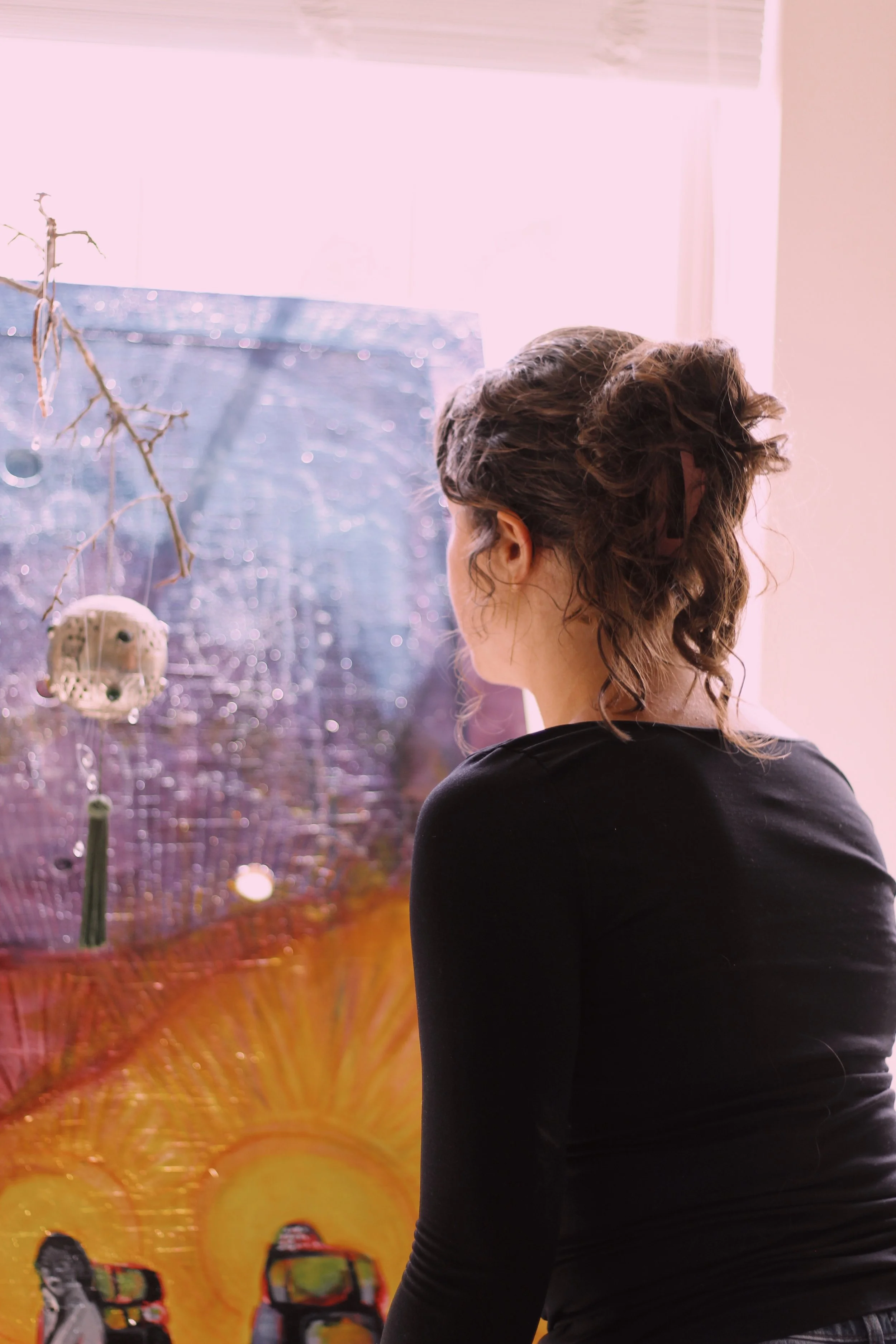 A person with curly hair wearing a black shirt is standing in front of a vibrant backdrop with colorful imagery including a sun and what appears to be snowmobiles, and a Dreamcatcher hanging on the left.