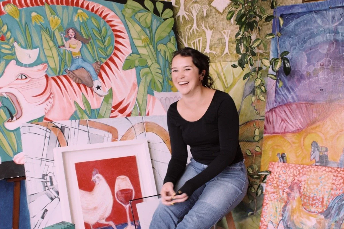 A woman sitting and smiling in an art gallery surrounded by colorful paintings, including a large mural of a woman on a tiger with greenery, a painting of a rooster with a glass of red wine, and other animal-themed artworks.