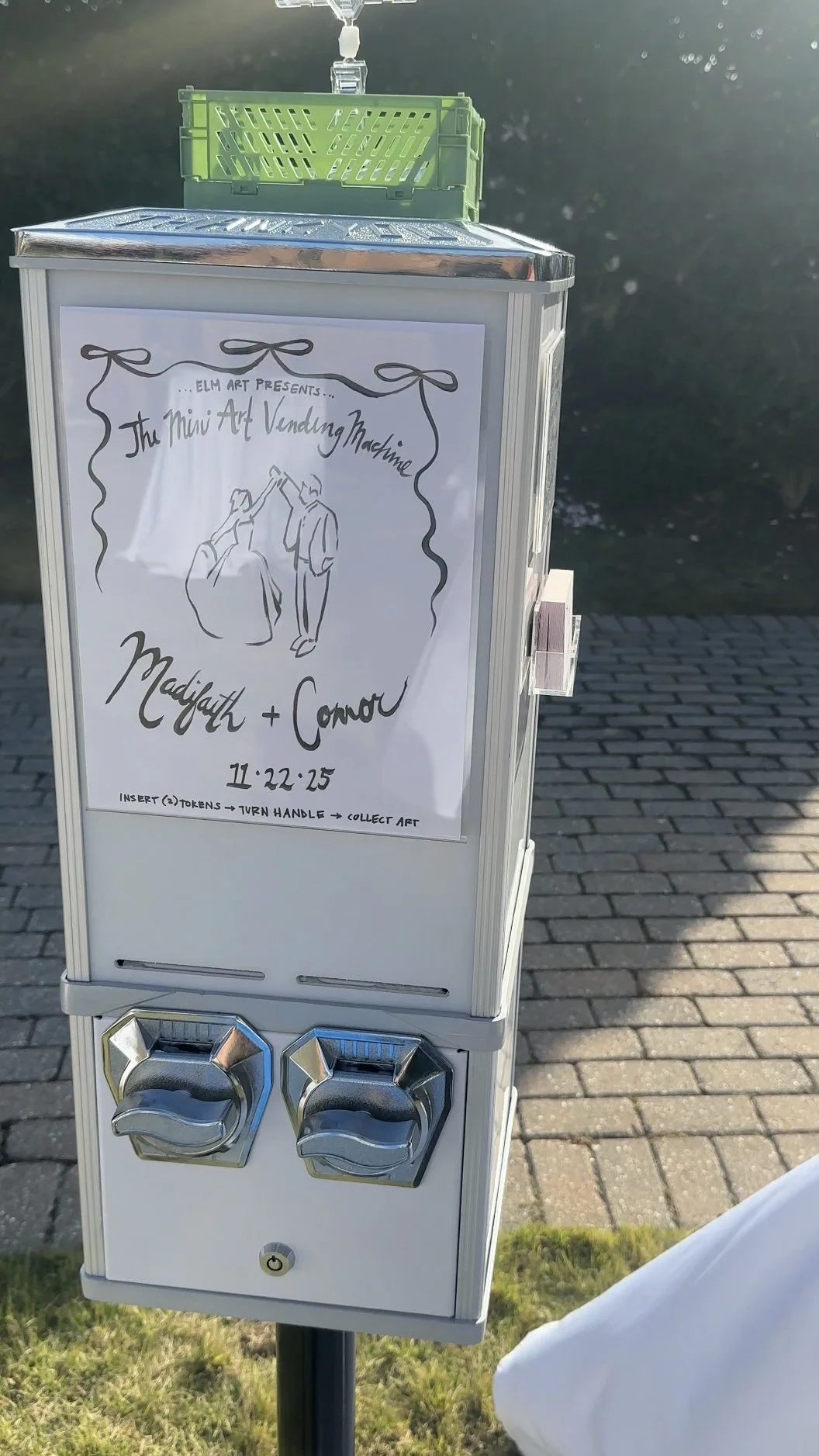 A vending machine for mini art sculptures with a sign reading 'Elm Art Presents... The Mini Art Vending Machine' featuring an illustration of two people exchanging artwork, the names Madelyth and Conor, and the time 11:22:25. Instructions at the bott