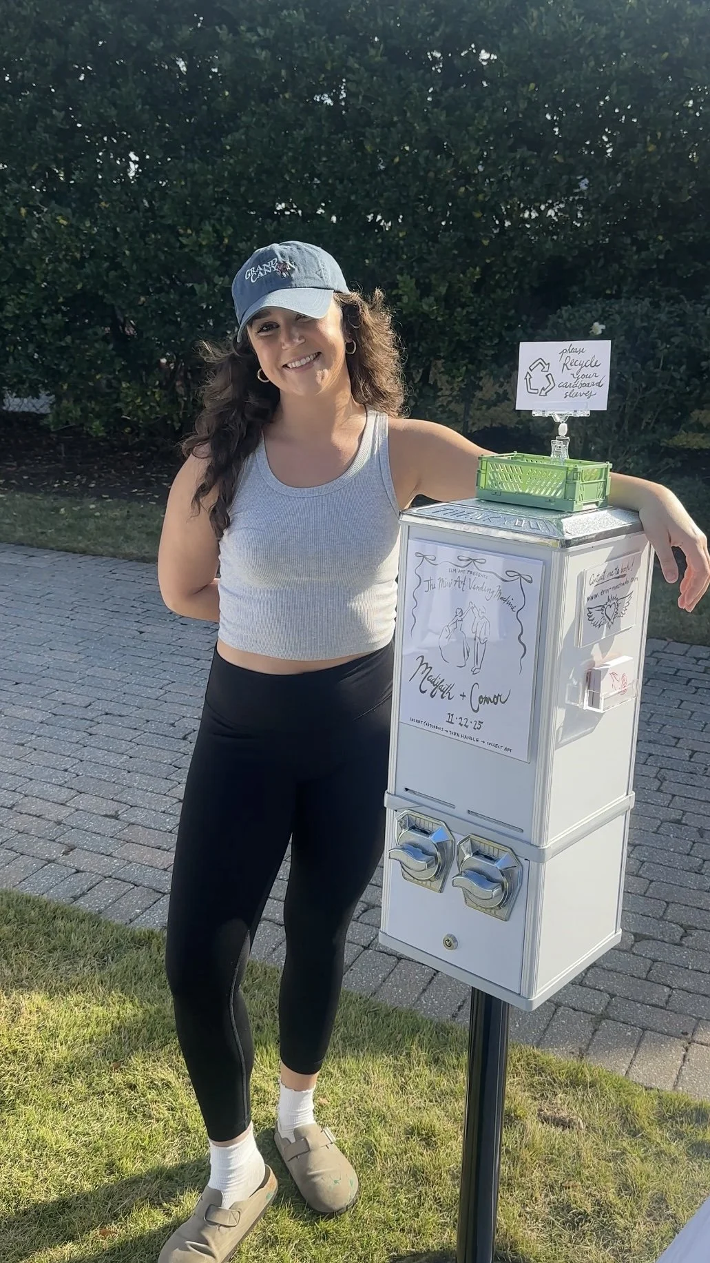 A smiling woman in a gray crop top, black leggings, and beige sandals stands next to a small vending machine for wedding donations, outdoors on a paved path with greenery behind her. The vending machine has a sign with a wedding illustration, and nex