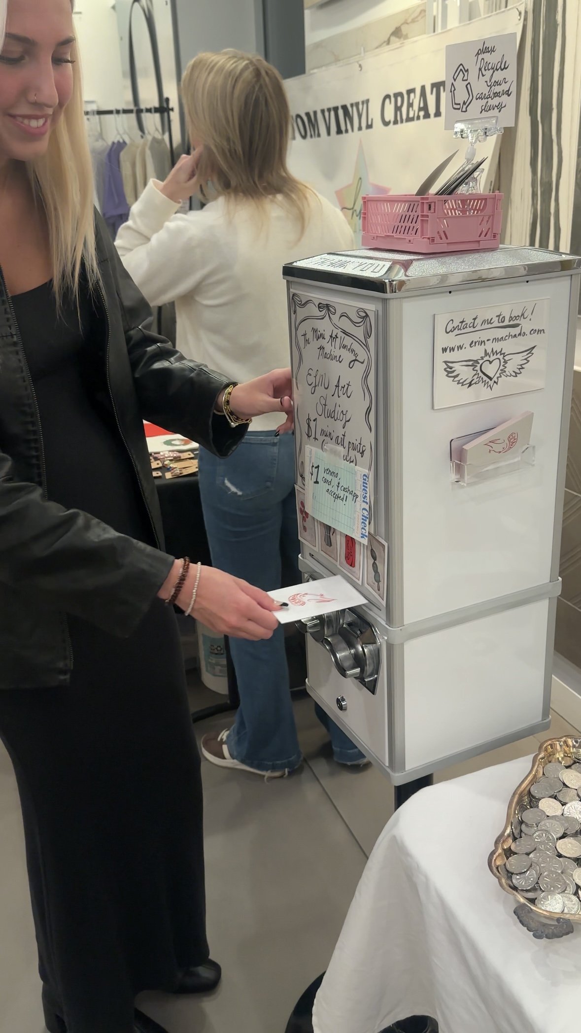 A woman is using a coin-operated machine to donate money, with a display of coins on a table nearby. The machine has signs indicating it is for a community art studio.