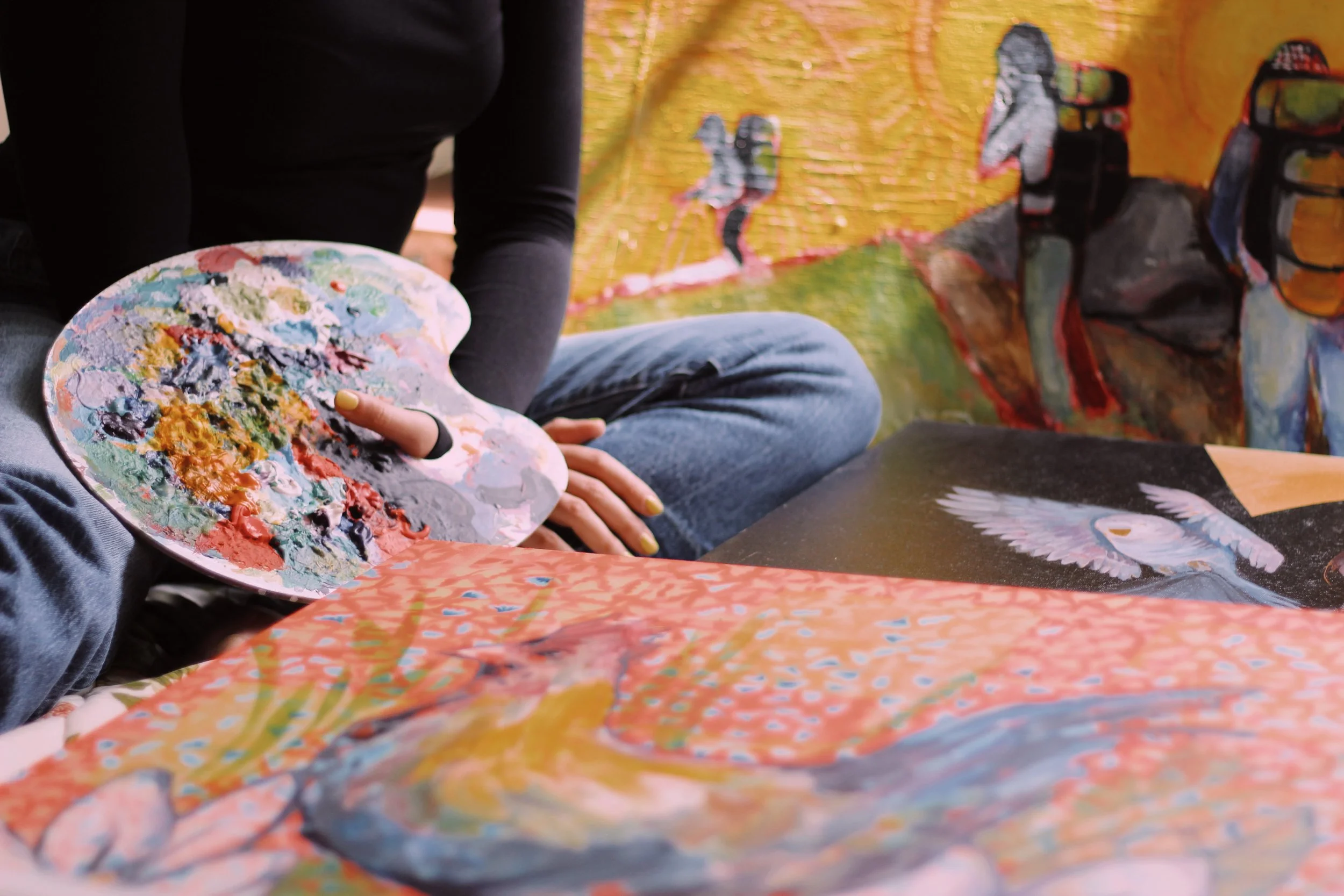 Person sitting on the floor with a paint palette, working on a colorful painting of a bird with outstretched wings.