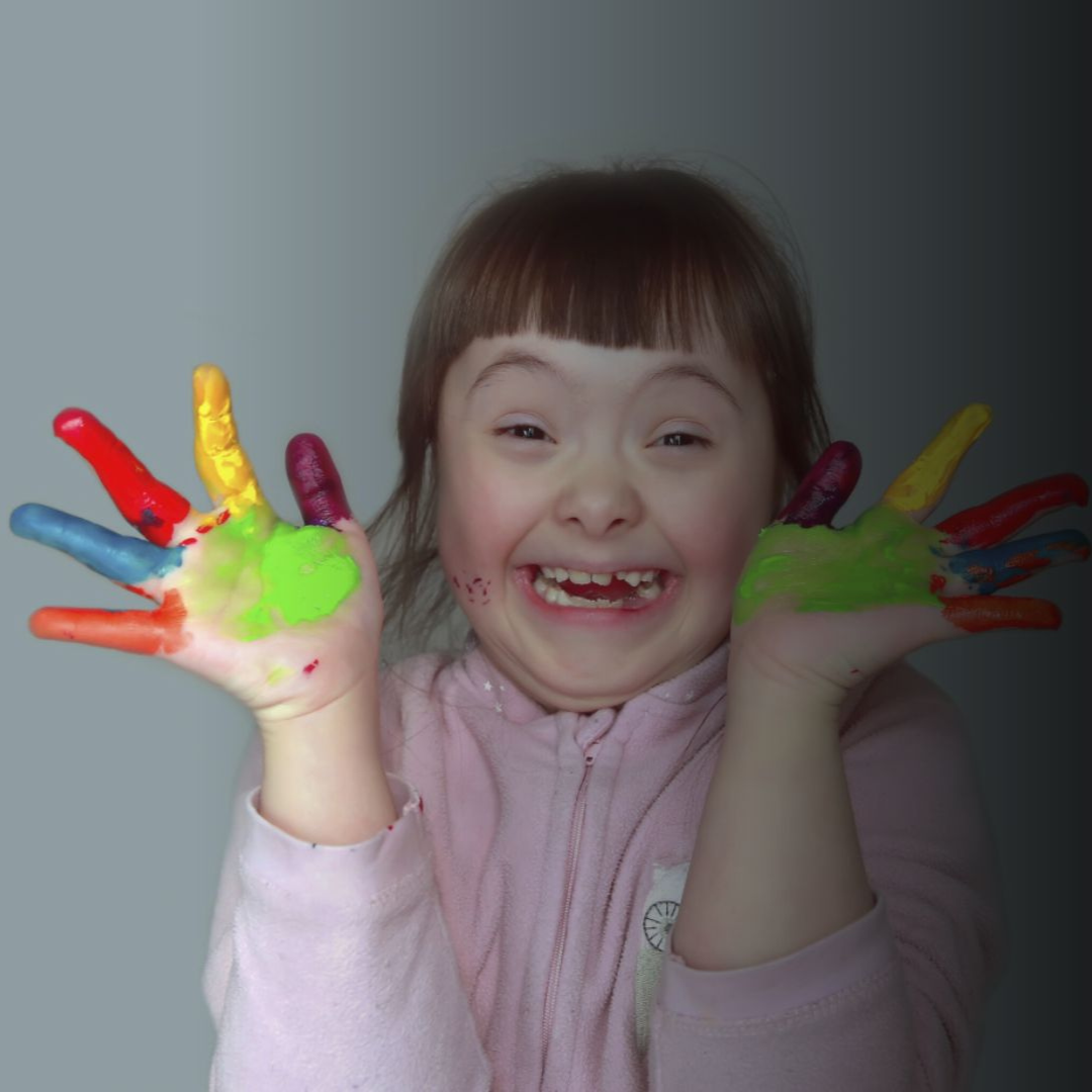 Niña sonriente con las manos cubiertas de pintura de varios colores y mostrando sus manos pintadas.