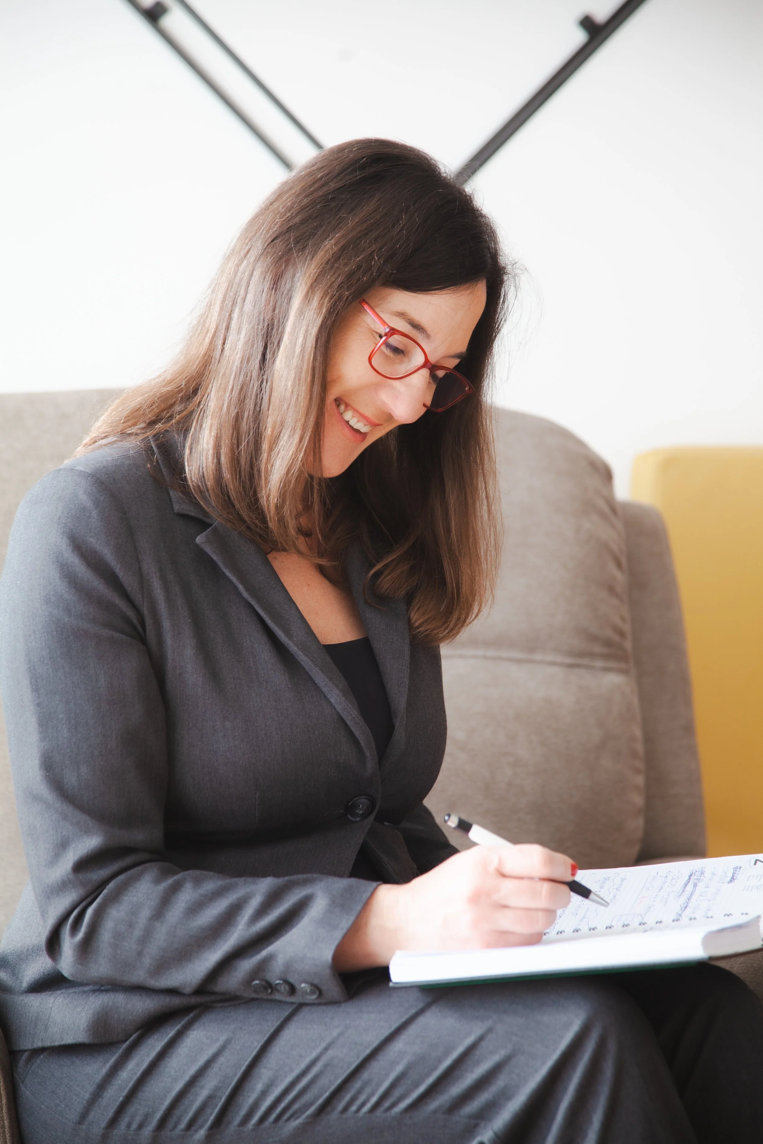 Woman in formal suit and red glasses sitting on a beige couch, writing notes in a notebook.