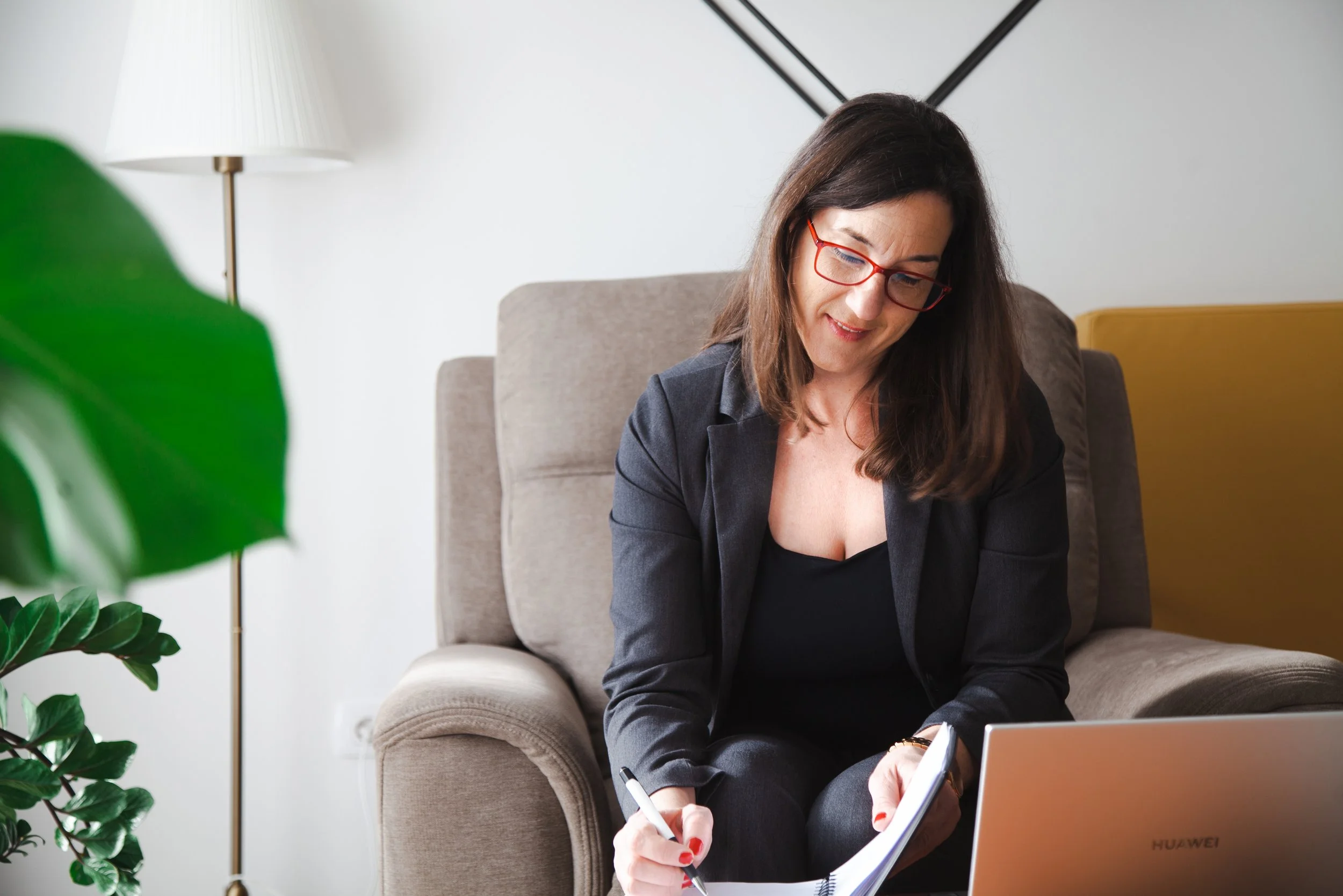 A woman with dark brown hair, wearing red glasses and a black blazer, sitting on a beige armchair, writing in a notebook, with a laptop in front of her and a green plant on her left.