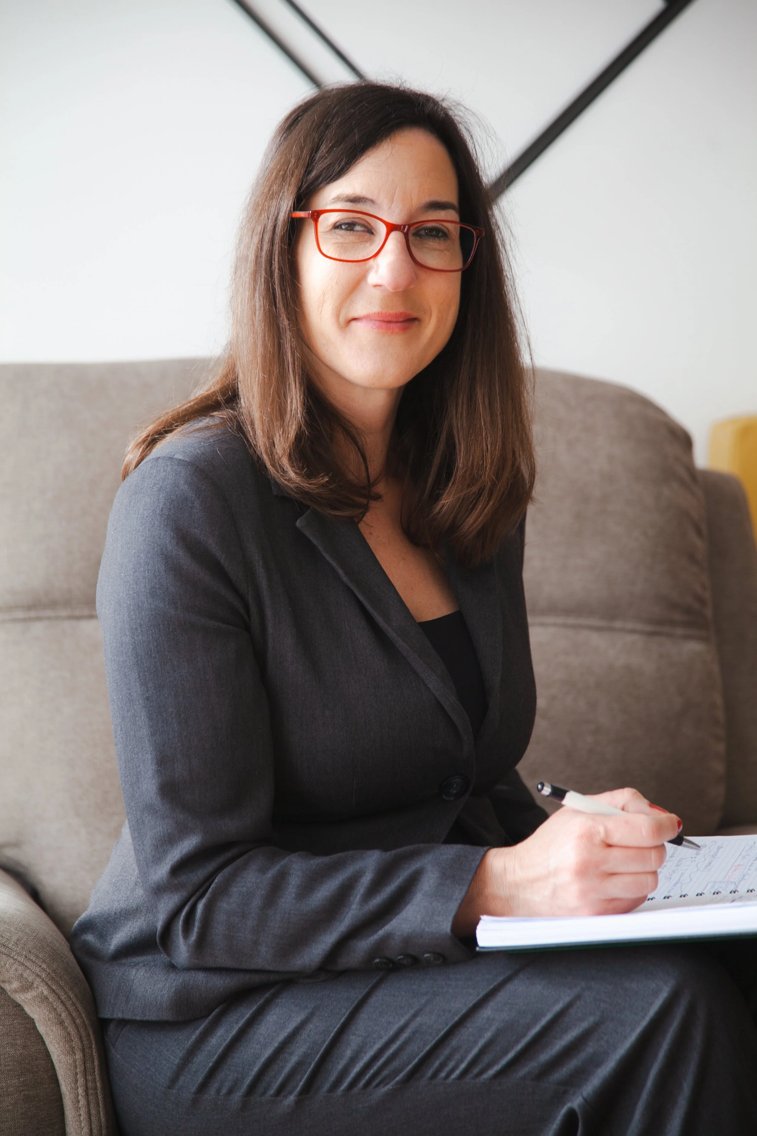 A woman with shoulder-length brown hair wearing red glasses and a dark gray business suit, sitting on a beige sofa, holding a pen and writing in a notepad, smiling at the camera.