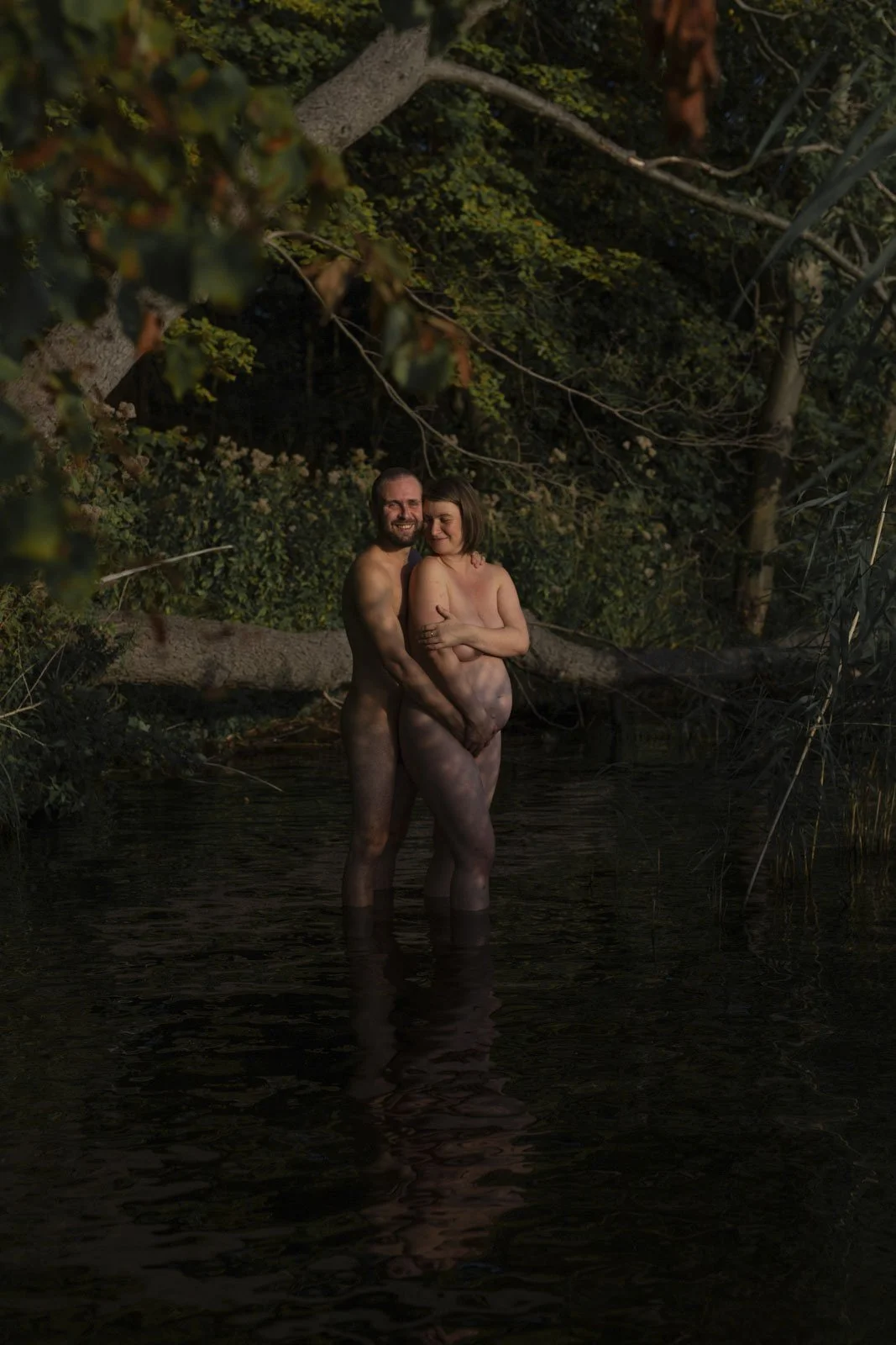 Ein Mann und eine Frau, nackt, umarmen sich in einem See im Wald, umgeben von Bäumen.