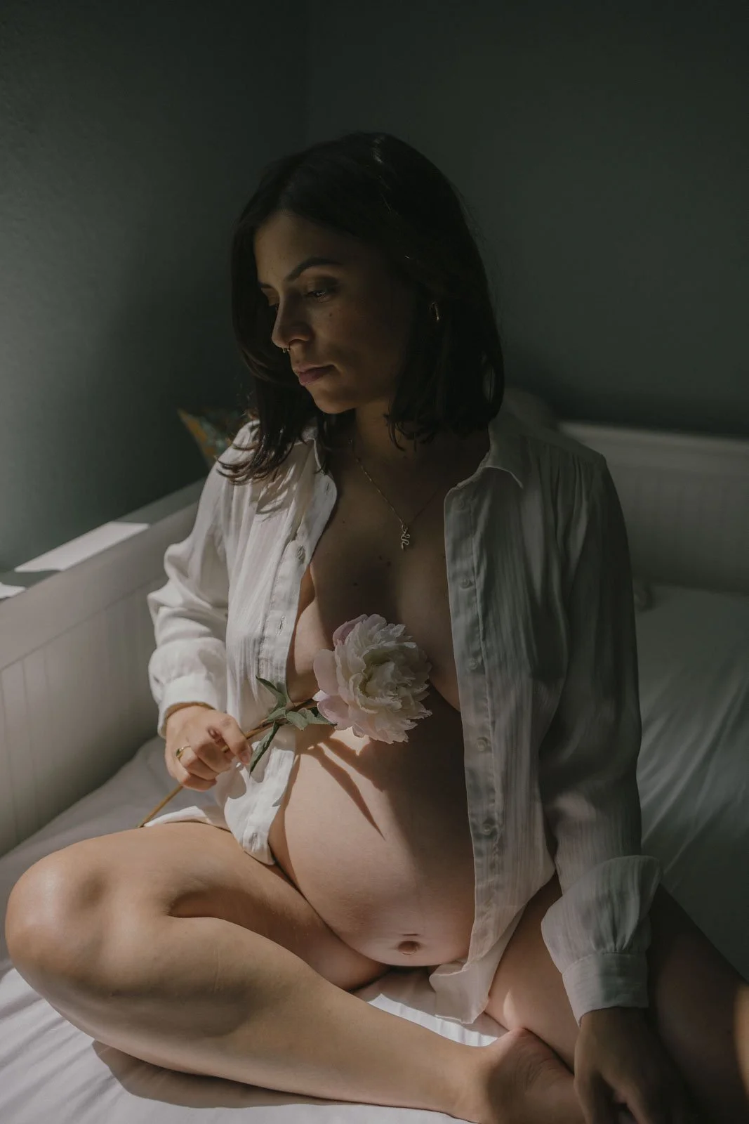 Eine schwangere Frau sitzt auf einem Bett, trägt eine offene weiße Bluse und hält eine große rosa Blume, der Raum ist dunkel und die Beleuchtung ist weich.