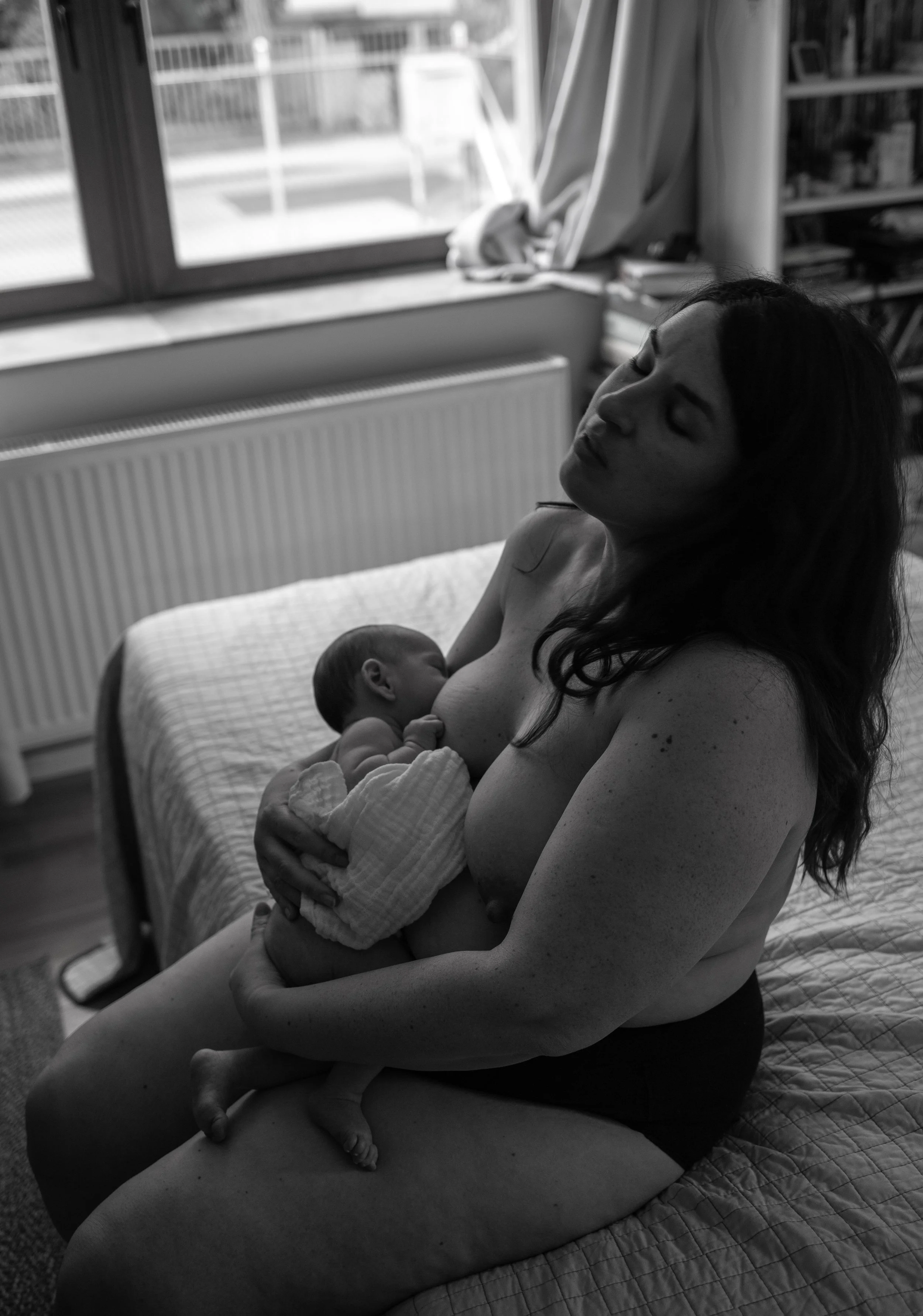 Schwarzweiß-Foto einer Frau, die auf einem Bett sitzt und ein Baby stillt, im Hintergrund ein Fenster und Bücherregale.