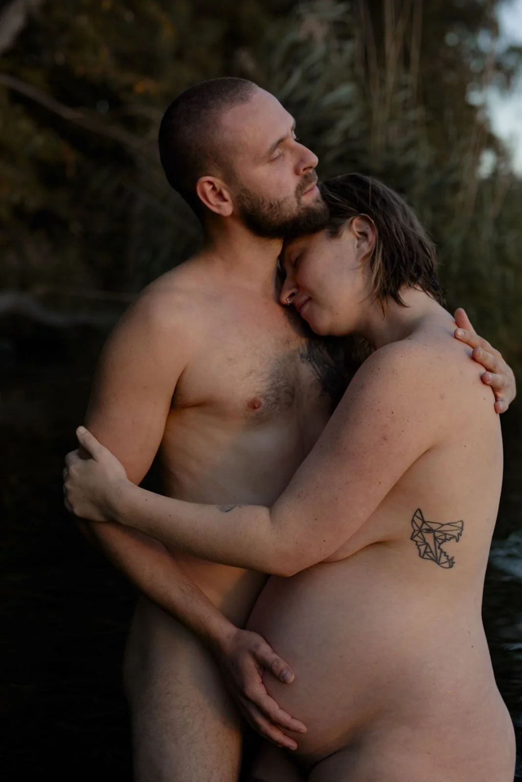 Ein nackt ein Paar steht im Wasser und umarmt sich liebkosend, umgeben von Natur. Der Mann hat einen Bart und eine Glatze, die Frau hat kurze Haare und ein Tattoo am Arm.