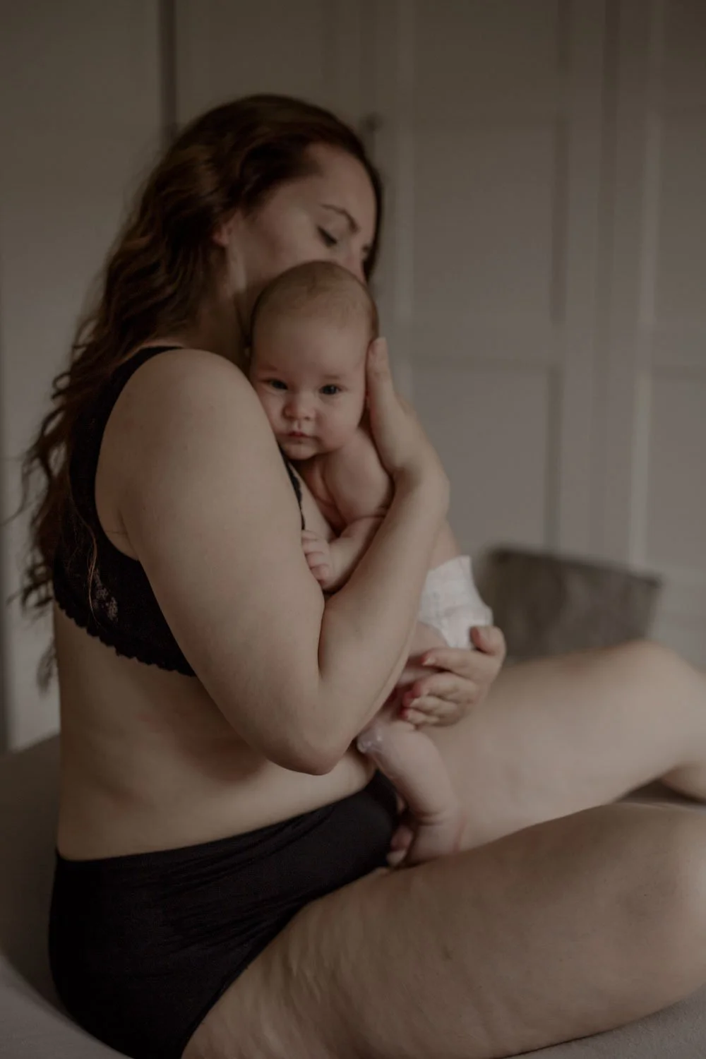 Eine Frau hält ein Baby in den Armen, das sich an ihre Schulter lehnt, in einem Raum mit weißem Wandpaneel, intime und ruhige Stimmung.