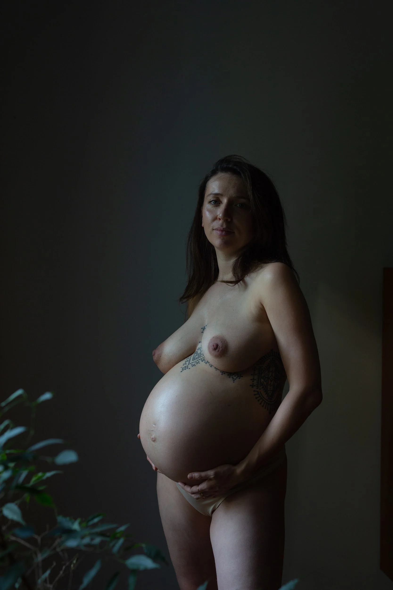 Schwangere nackte Frau mit Tattoos posiert in einem Raum mit dunklem Hintergrund, neben einer Pflanze.